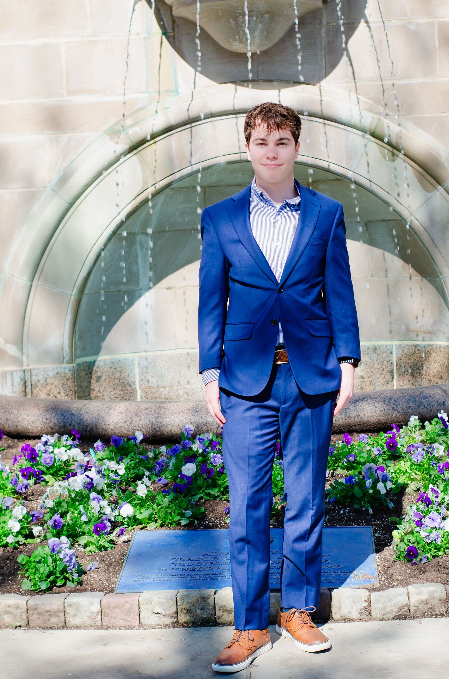 Young man in blue suit posing confidently in front of a stone fountain with colorful purple and white flowers.
