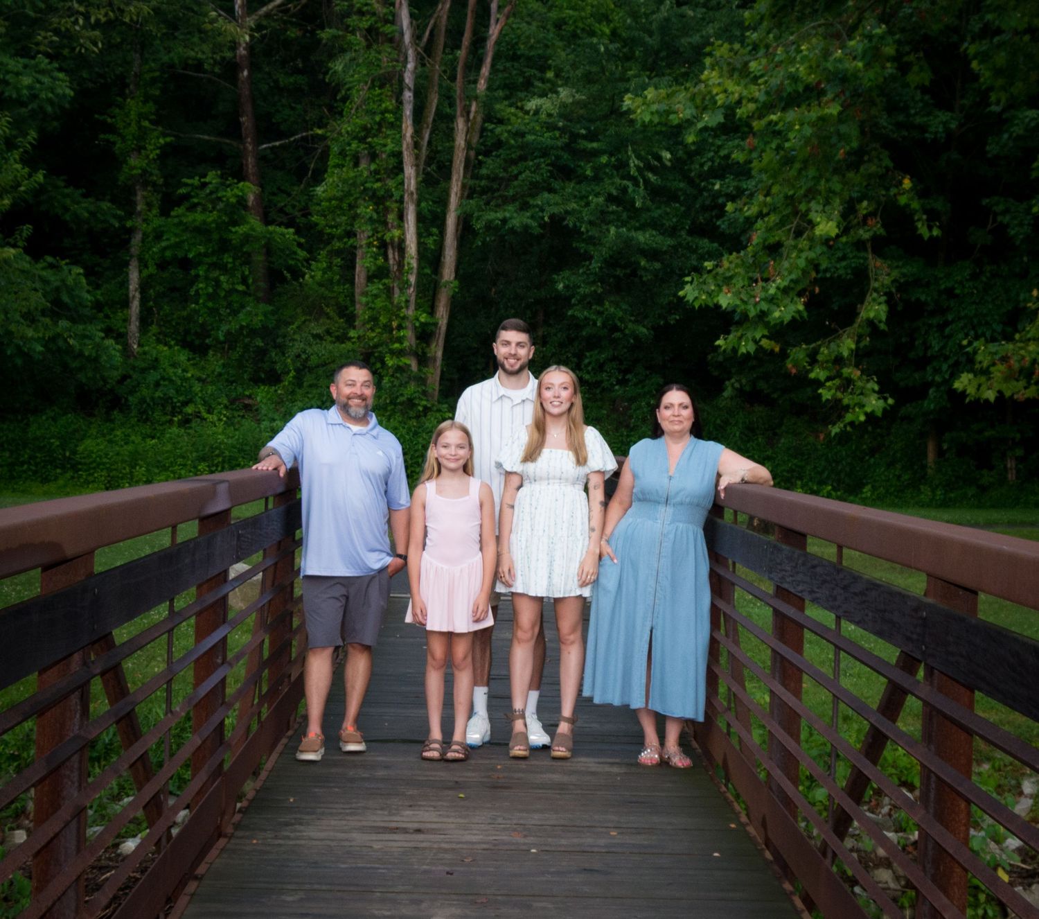 Family group poses together on wooden bridge in lush green park setting.