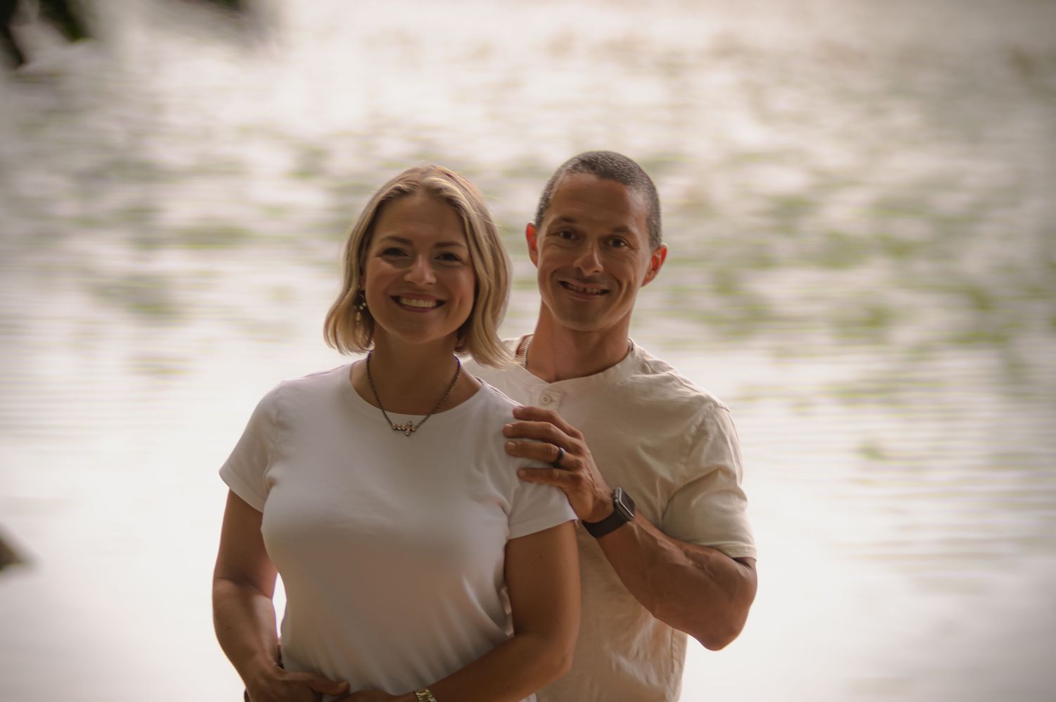 Happy couple embraces while standing near a reflective lake on a bright summer day.