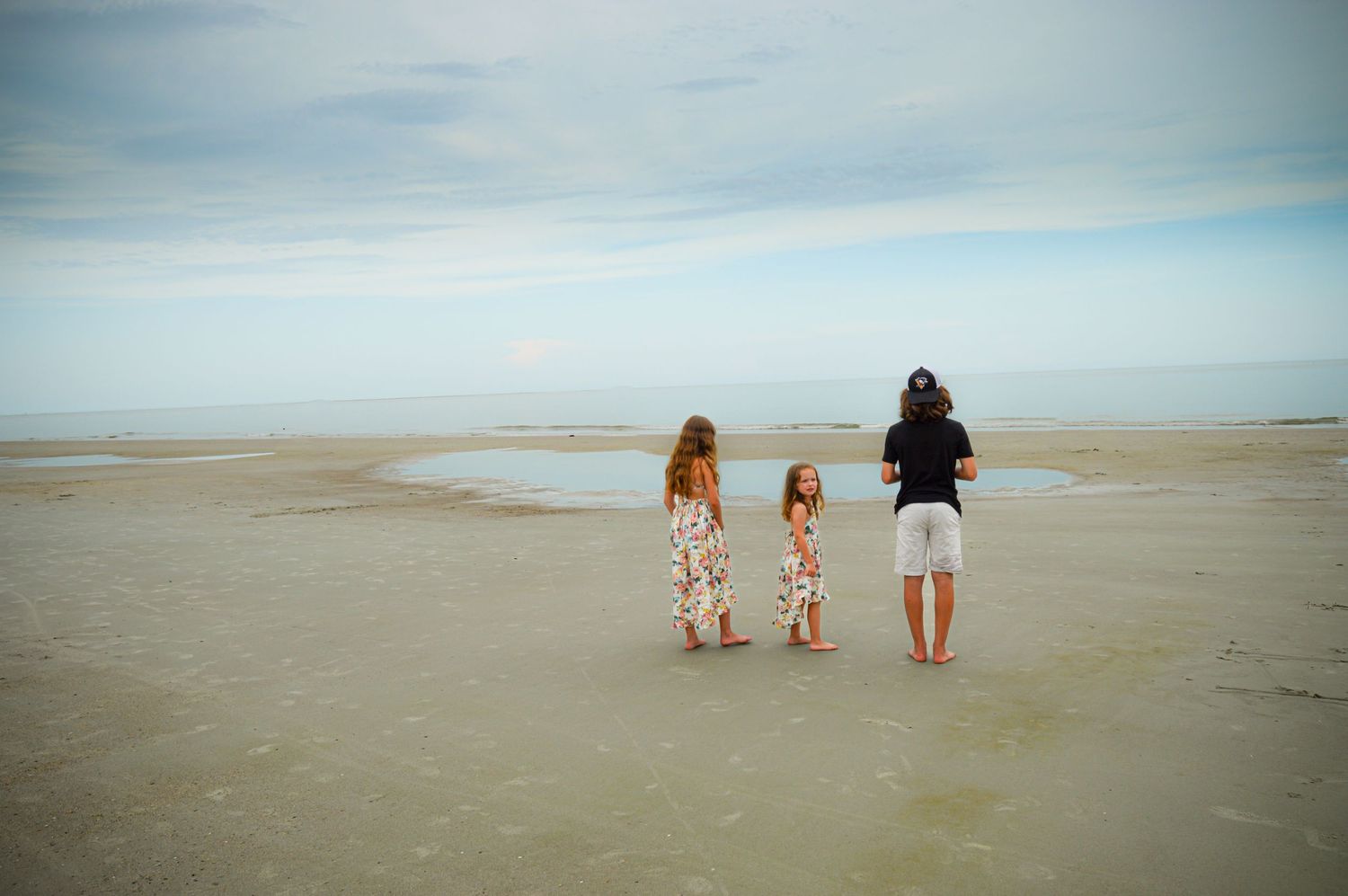 Three people stand looking out at the ocean on a wide sandy beach.