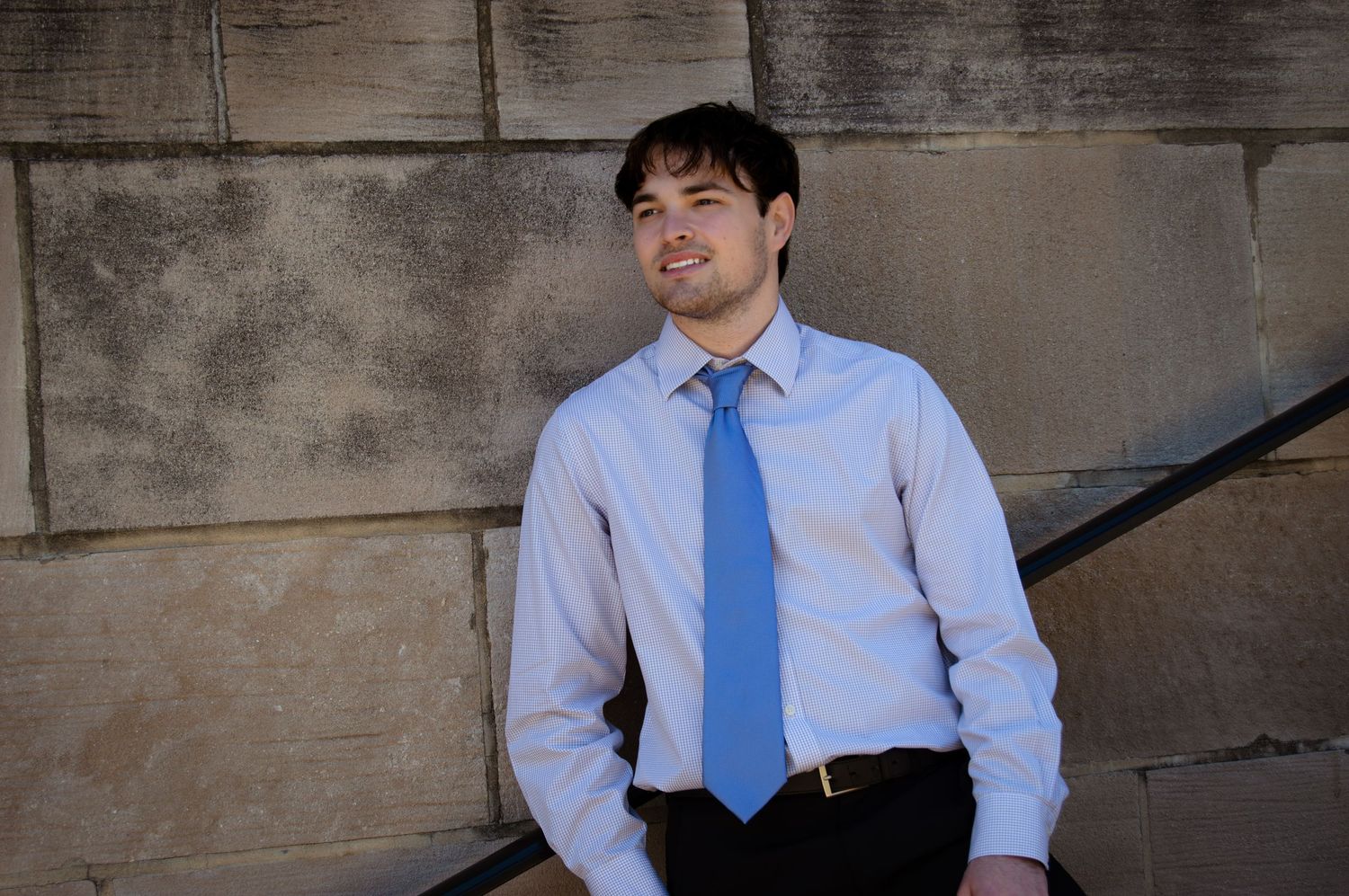 Young man in blue dress shirt and tie leaning against stone wall, smiling outdoors.