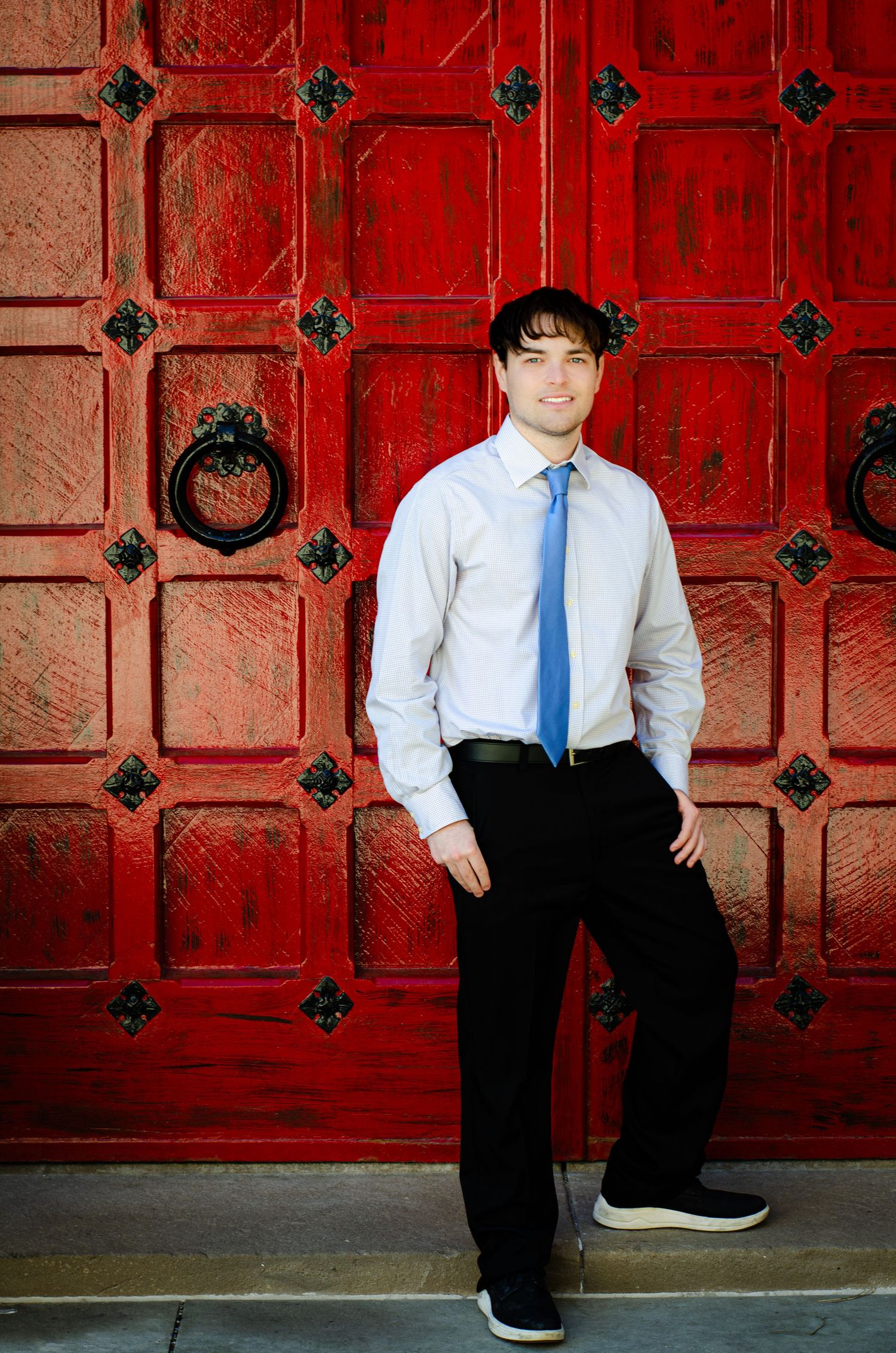 Young man in blue tie and white shirt posing confidently against ornate red wooden doors with black iron hardware.