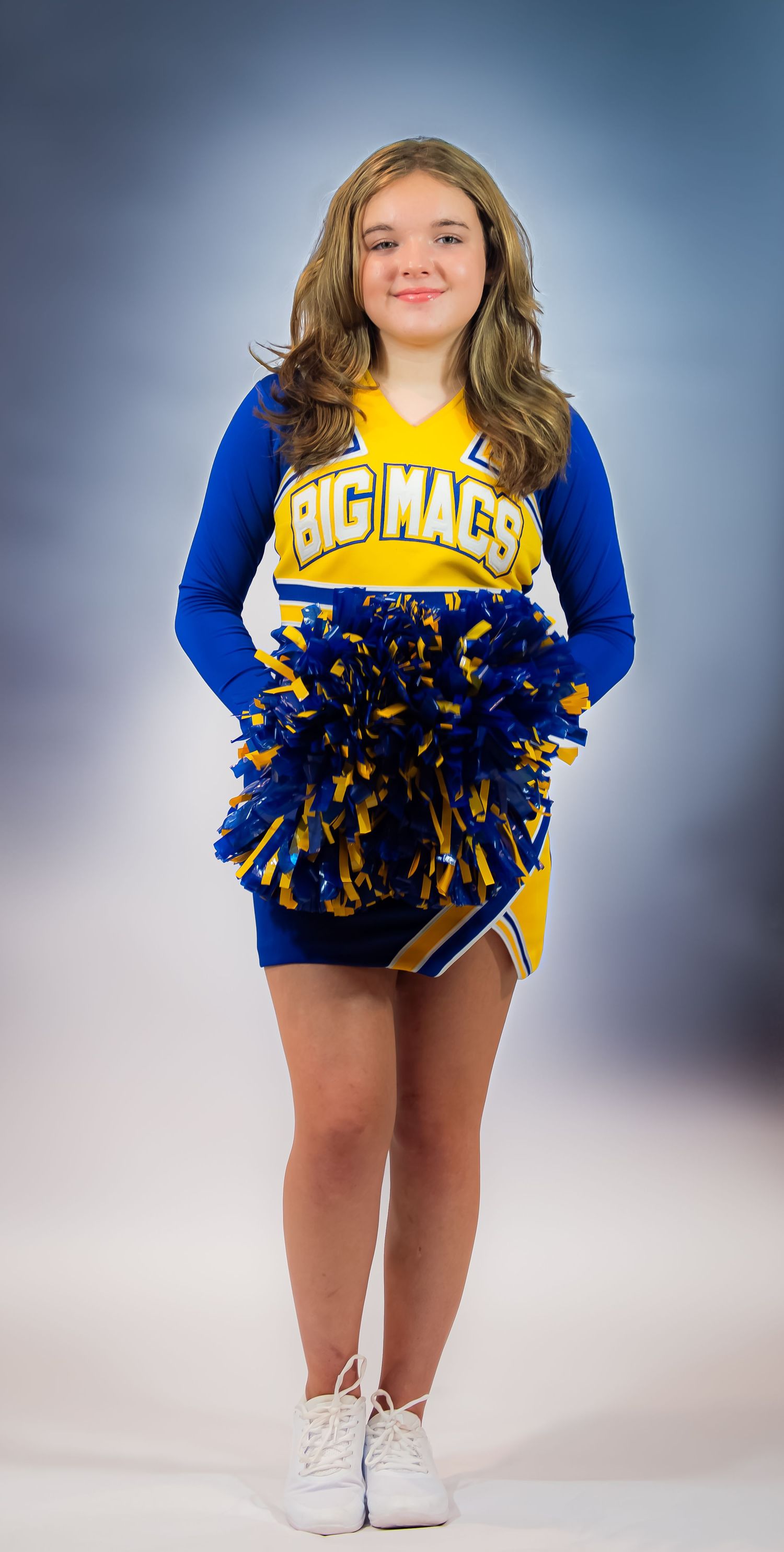 A cheerleader in blue and gold uniform poses with pom poms against a blue studio background.