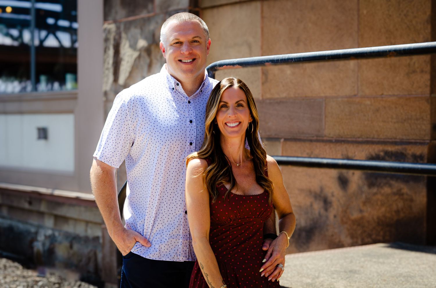 Smiling couple posing together outdoors against a stone wall on a sunny day.