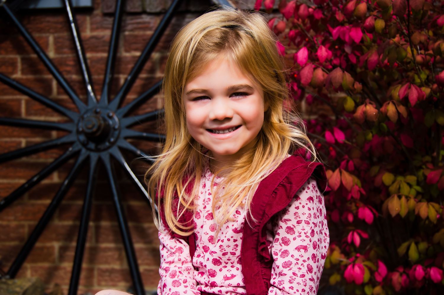 A young child in a pink floral dress and burgundy vest poses outdoors against colorful autumn foliage.