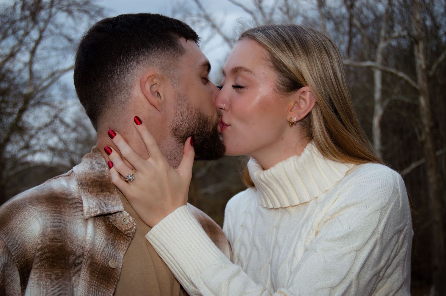 A romantic couple shares a tender kiss outdoors while wearing cozy winter sweaters on a cloudy day.