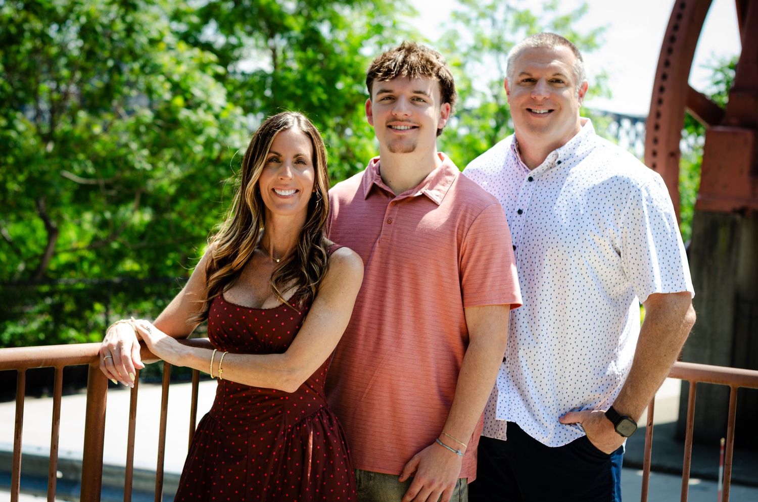 Three smiling people pose on a bridge surrounded by lush green trees in a warm outdoor family portrait.