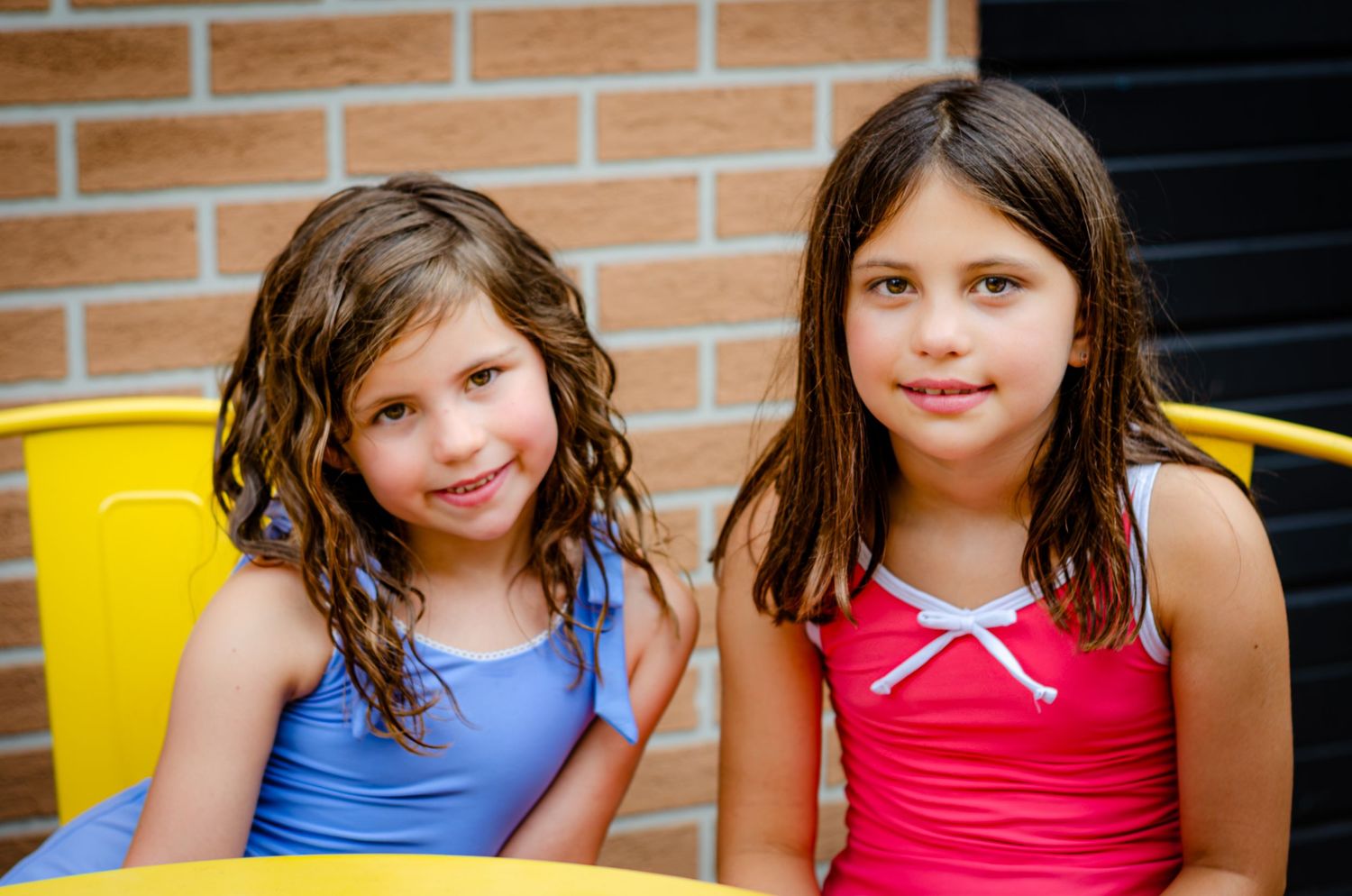 Two young girls with wet hair in colorful swimsuits smile while sitting in yellow chairs outdoors near a brick wall.