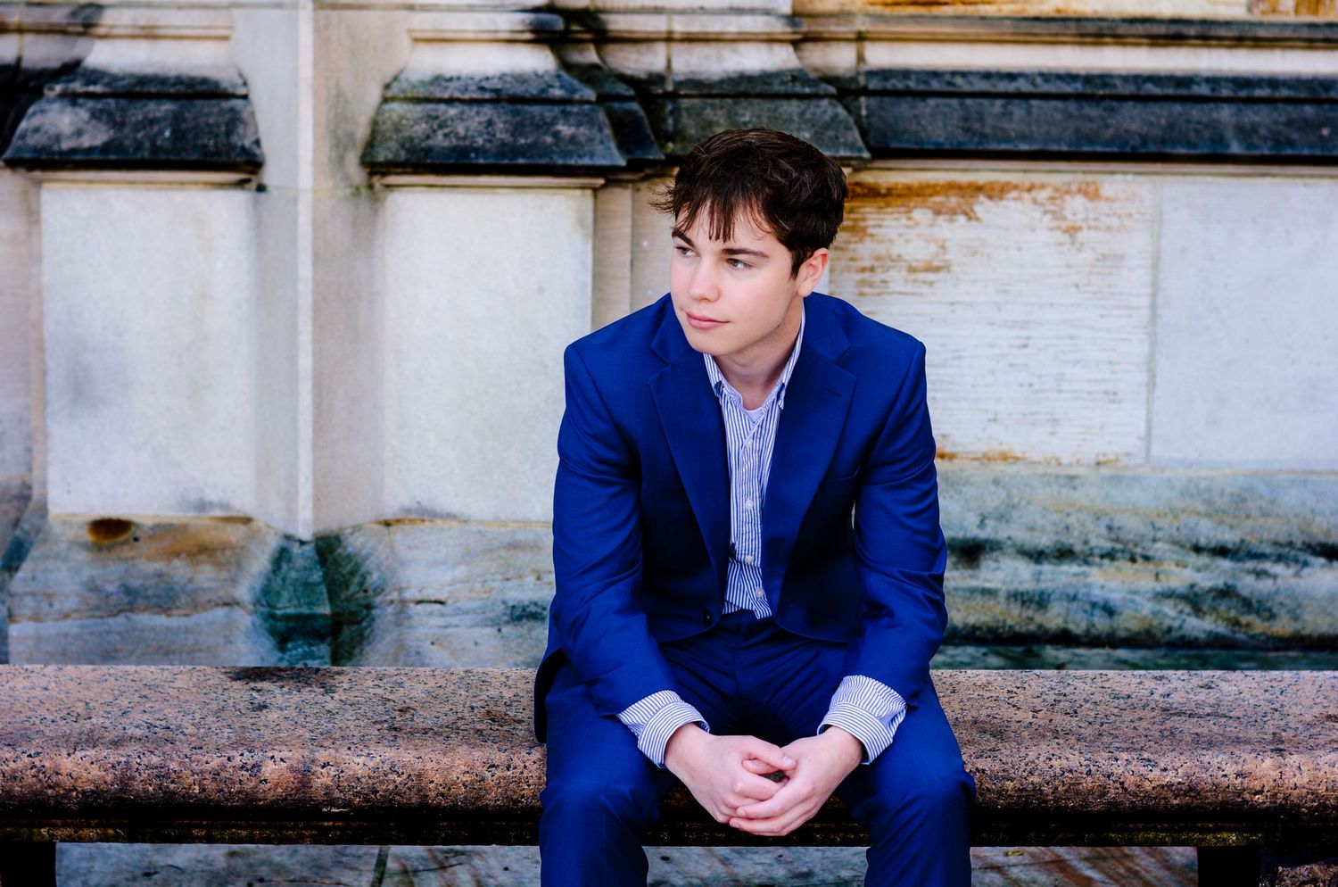 Young man in a blue suit sitting on stone steps outdoors, looking thoughtfully to the side.