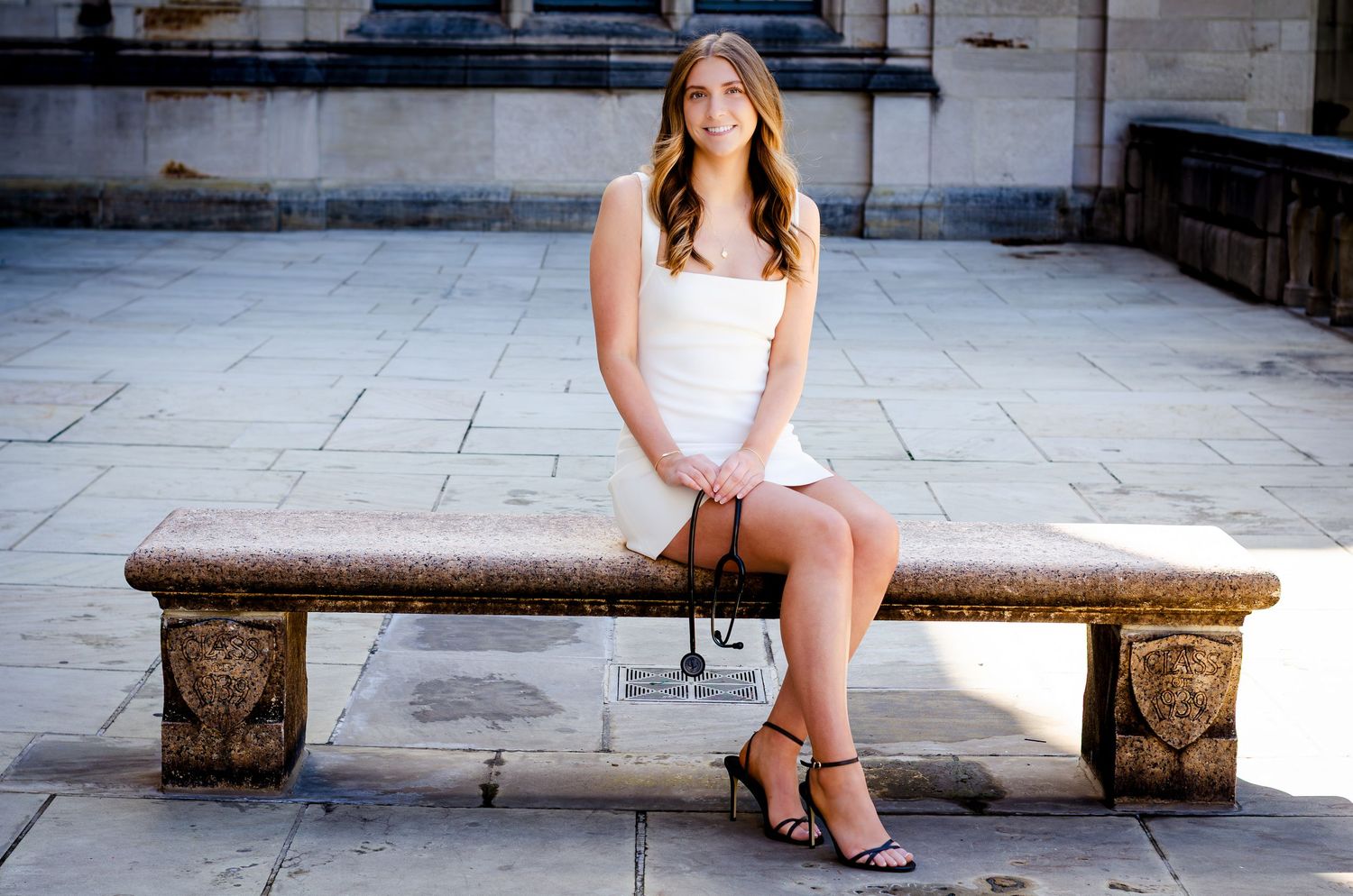 Young woman in white dress sitting on ornate stone bench outdoors smiling for senior portrait photo.