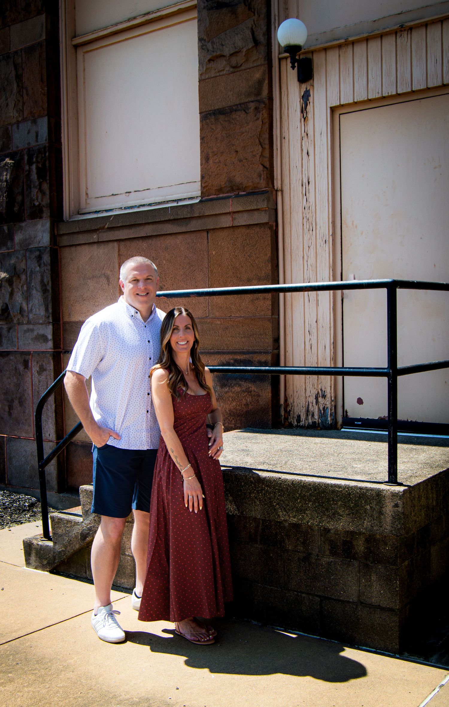 Couple smiling together on sunny day outside historic stone building with black metal railing.