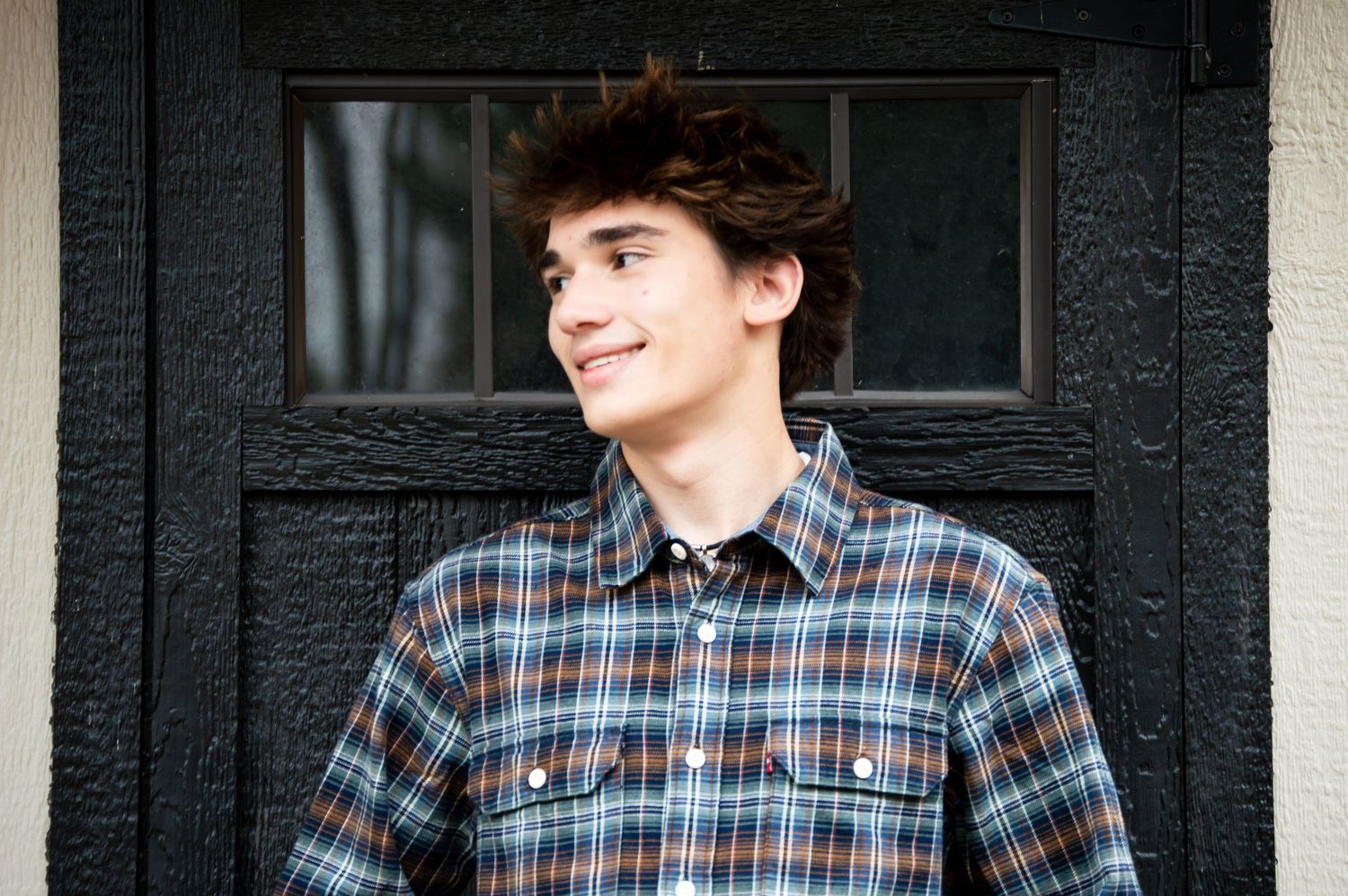 A person in a blue plaid shirt and denim poses casually by a dark door with dried grass in view.