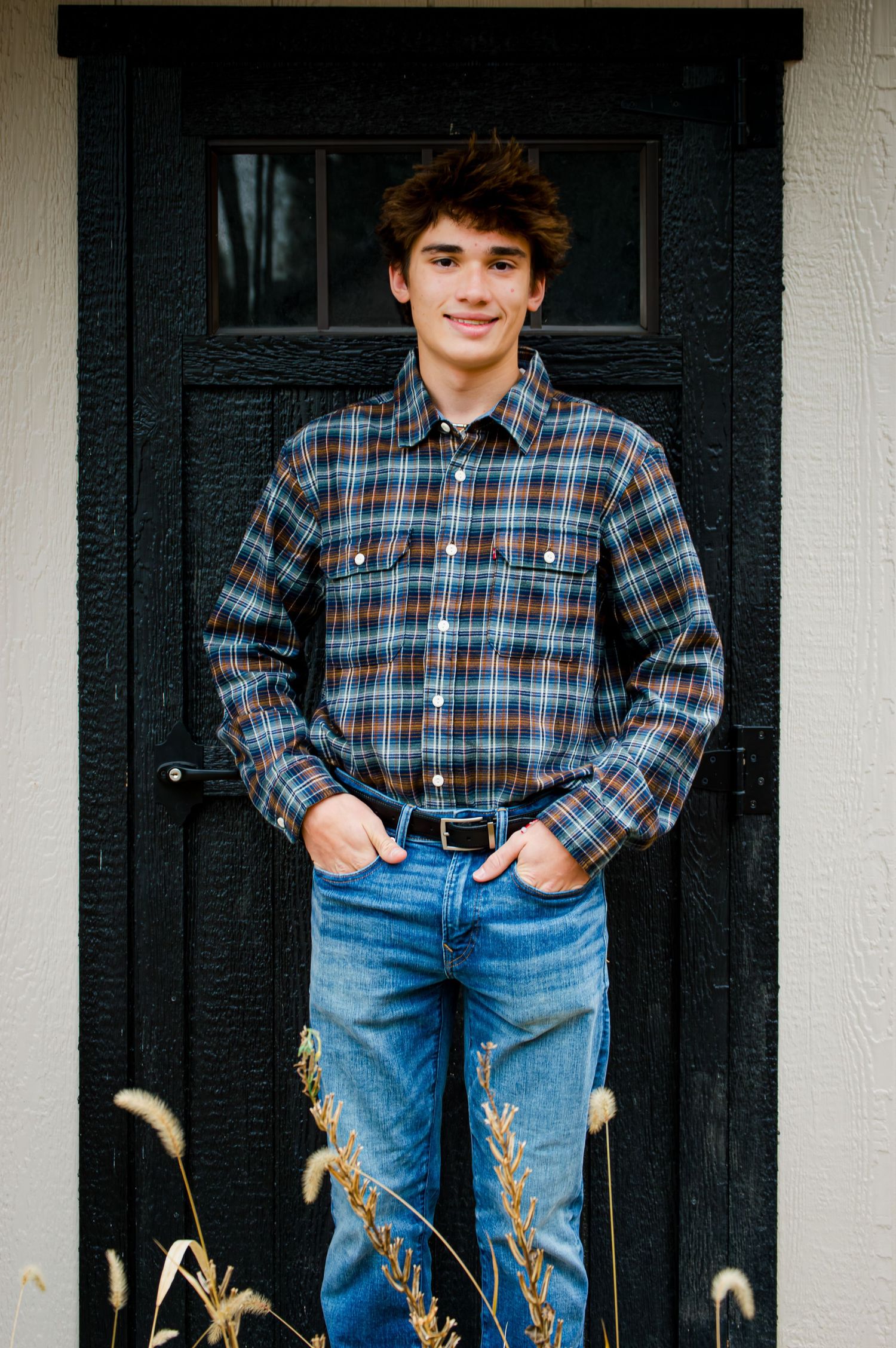 A person wearing a plaid flannel shirt and jeans stands against a black door with tall grass in the foreground.