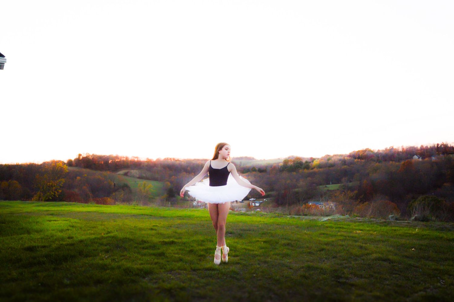 A person stands in a green meadow at sunset with a white farmhouse visible in the distance.