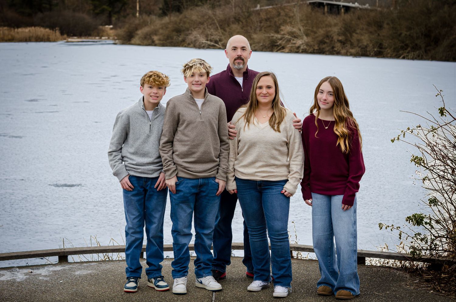 Family members stand together outdoors by a lake on an overcast day wearing coordinated casual outfits.