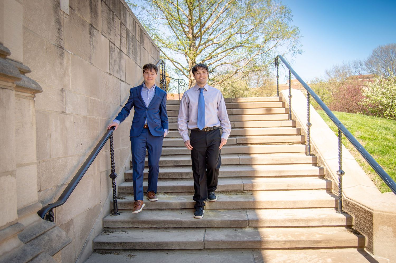 Two young men in suits walking down outdoor stone steps on sunny day.