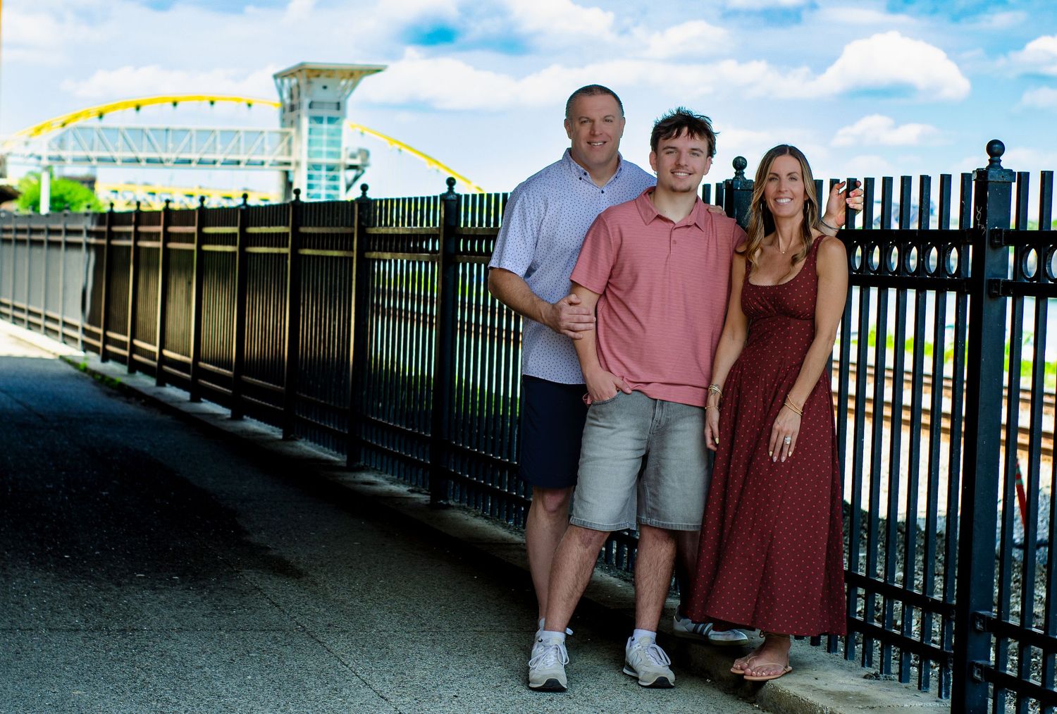 Three people smiling on a bridge walkway with a yellow arch bridge and stadium visible in the background.