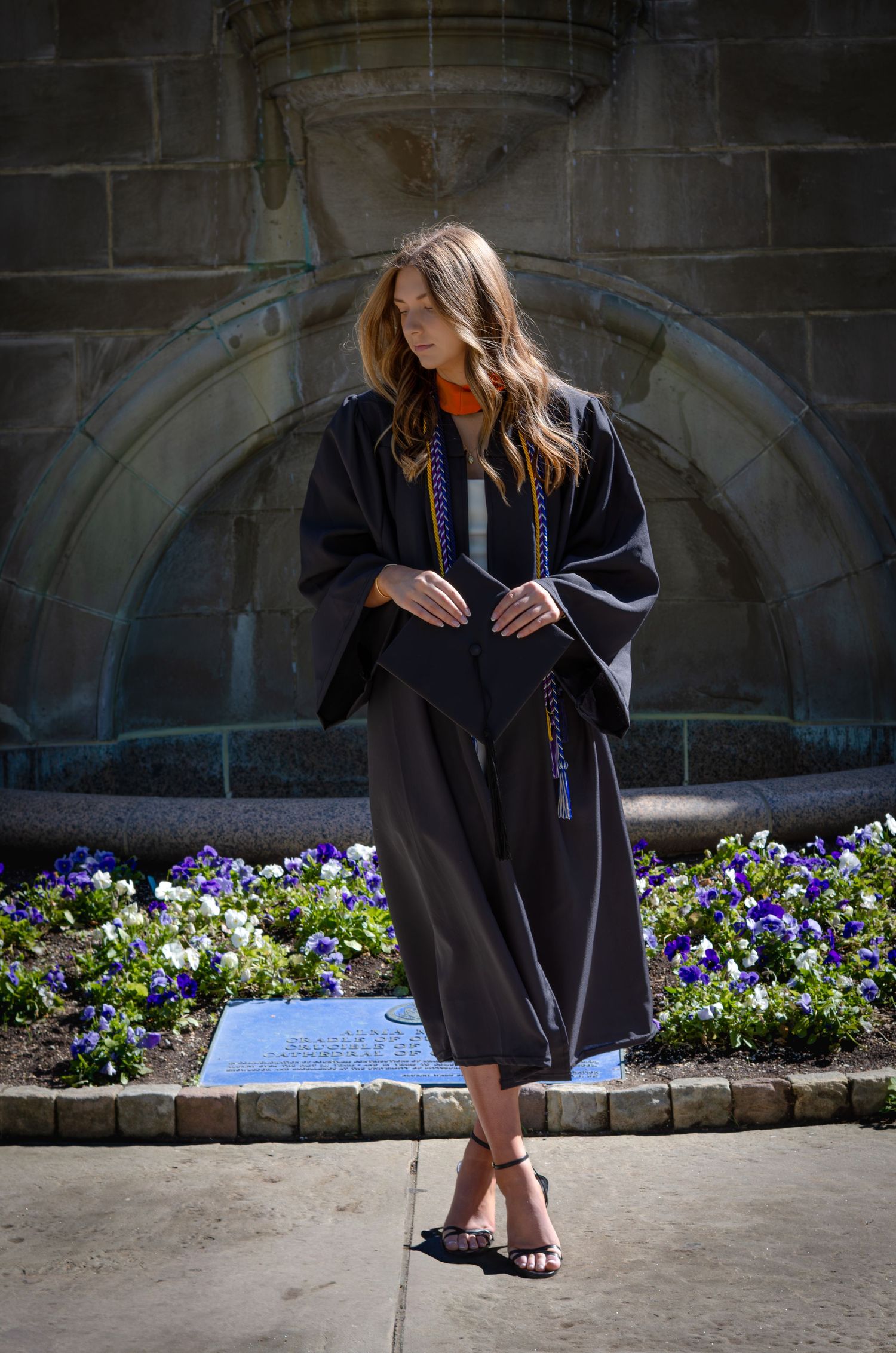 Young woman in black graduation gown and colorful honor cords stands before stone arch with purple flowers.