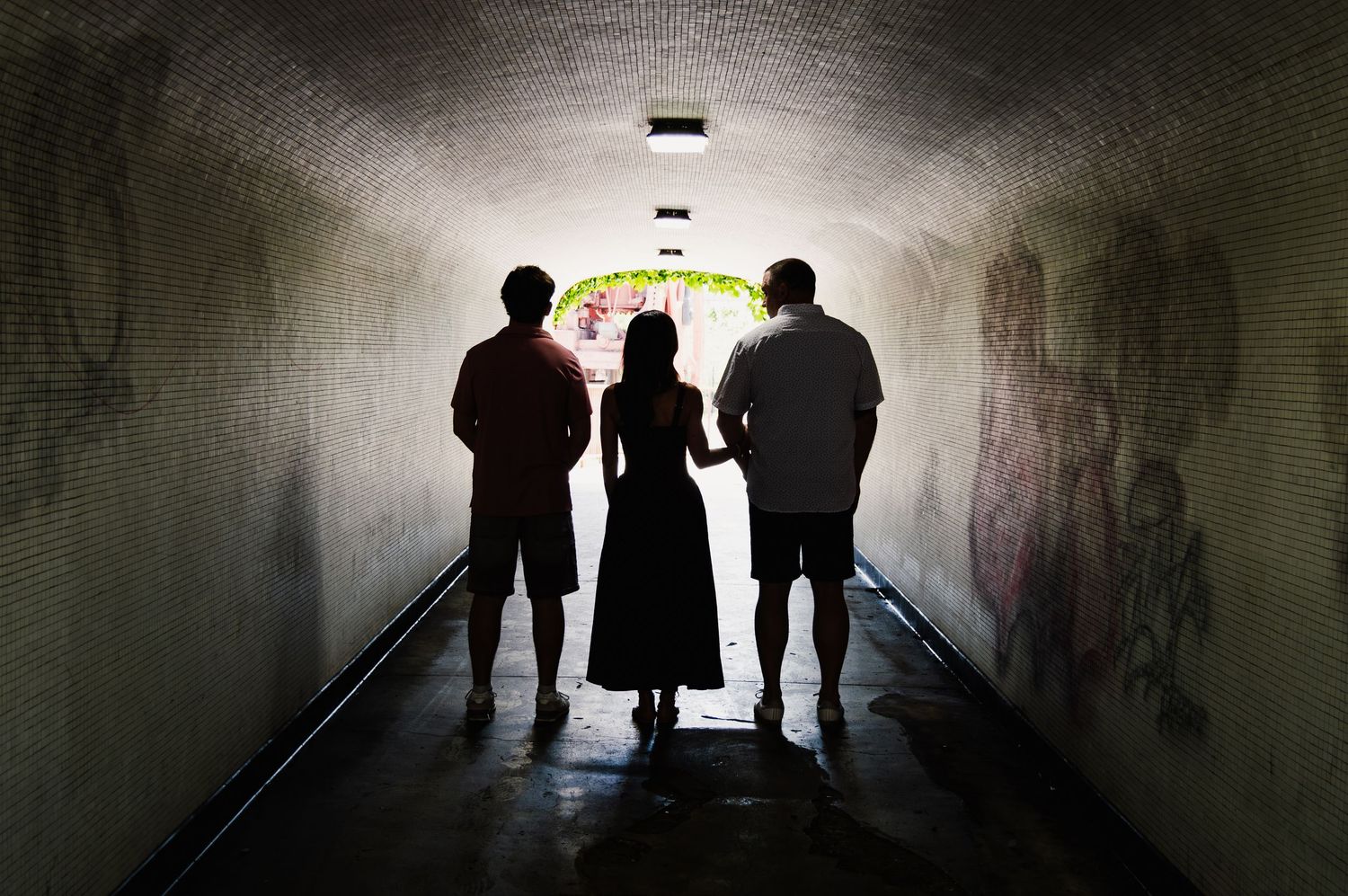 Three silhouetted figures stand in a graffiti-lined tunnel, gazing toward a bright, flower-adorned exit.