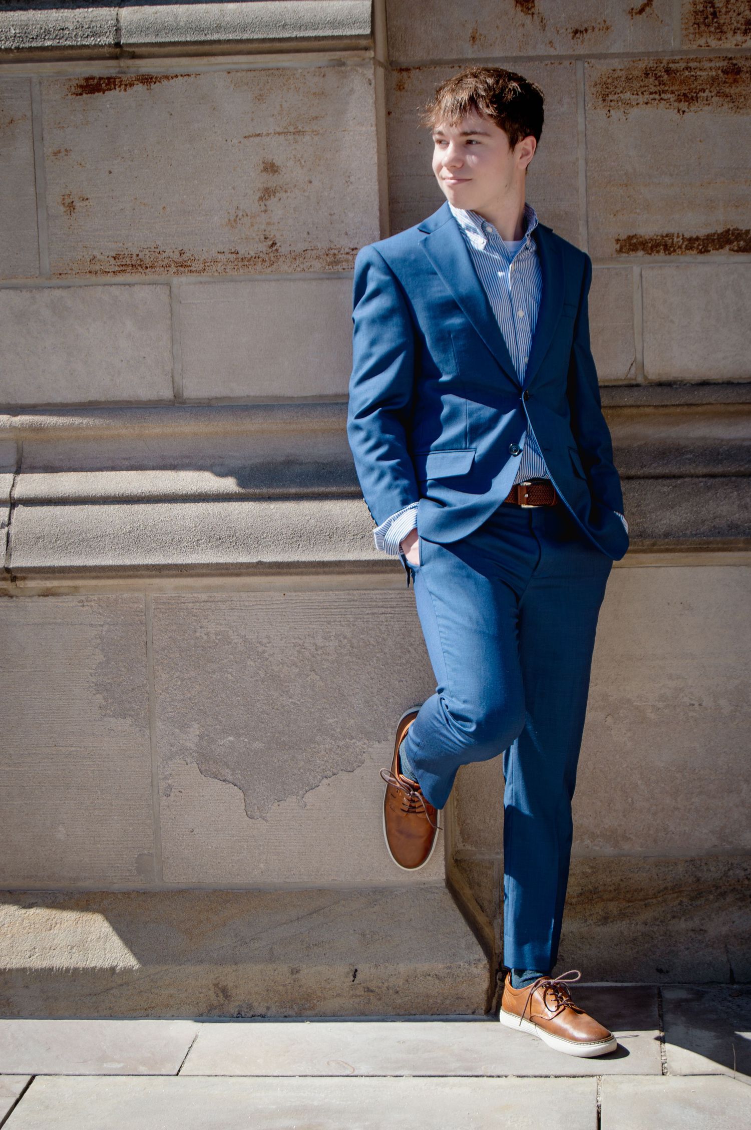 Young man in blue suit with brown shoes leaning against stone wall in sunlight.