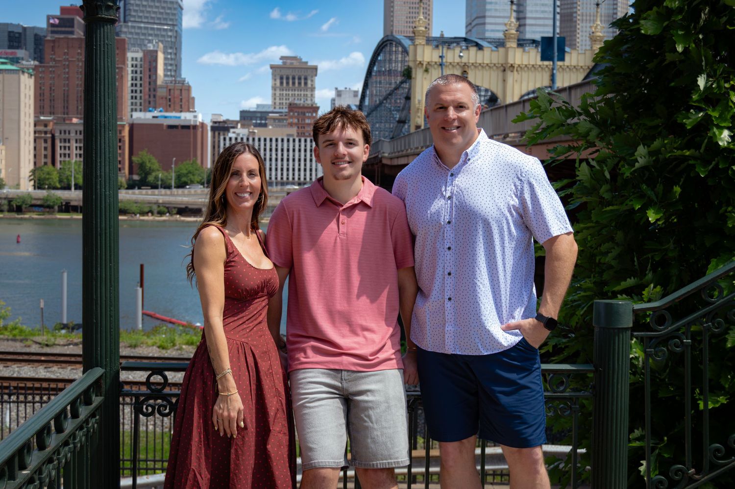 Family of three smiling with Pittsburgh city skyline and river in the background.