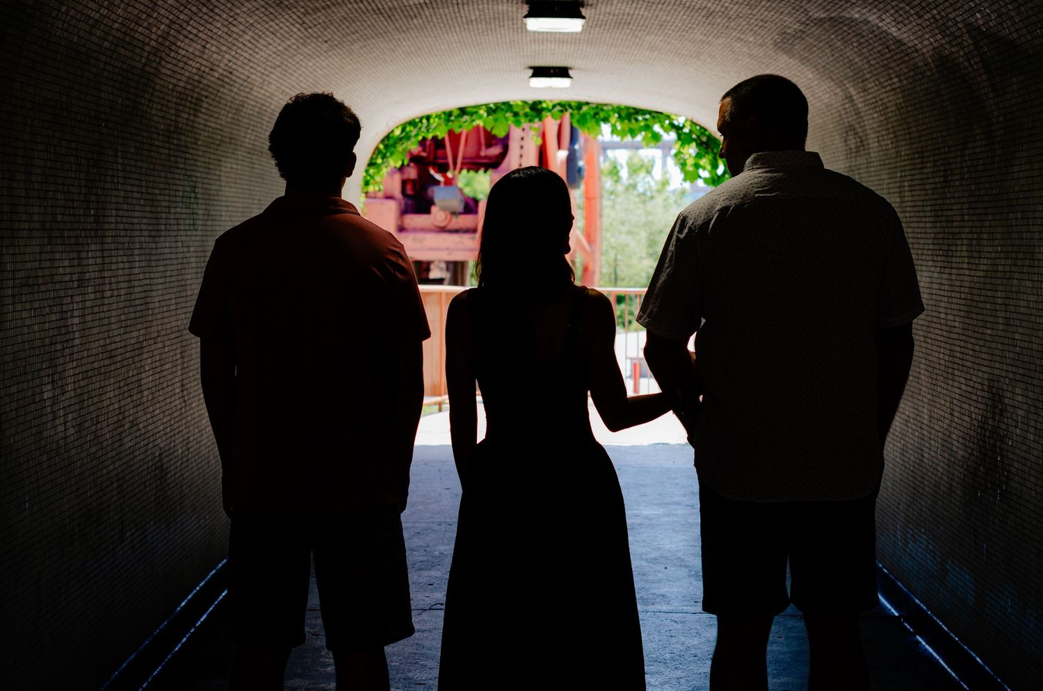 Silhouettes of three people standing in a tunnel archway with sunlit courtyard beyond.
