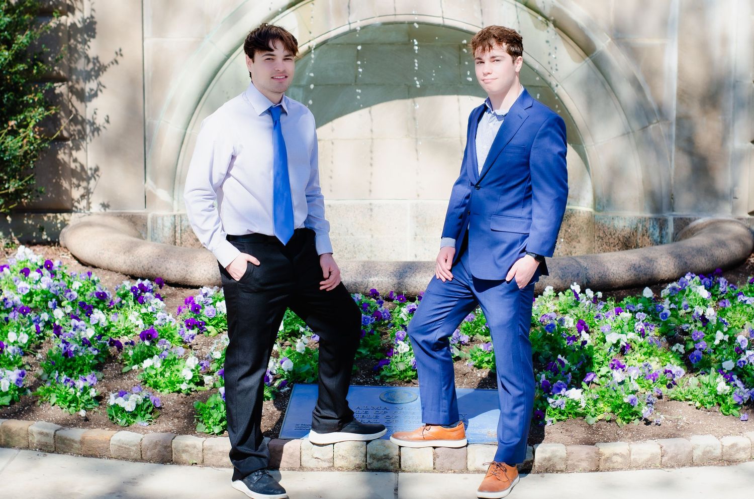 Two young men in dress attire posing near floral garden fountain outdoors.