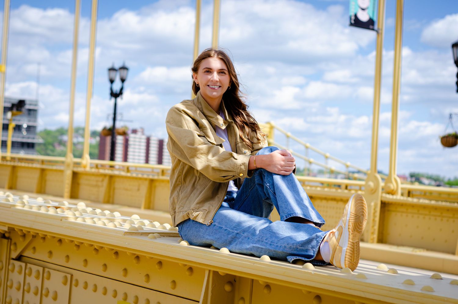 A young woman in jeans sits smiling on a yellow bridge with a blue sky backdrop.