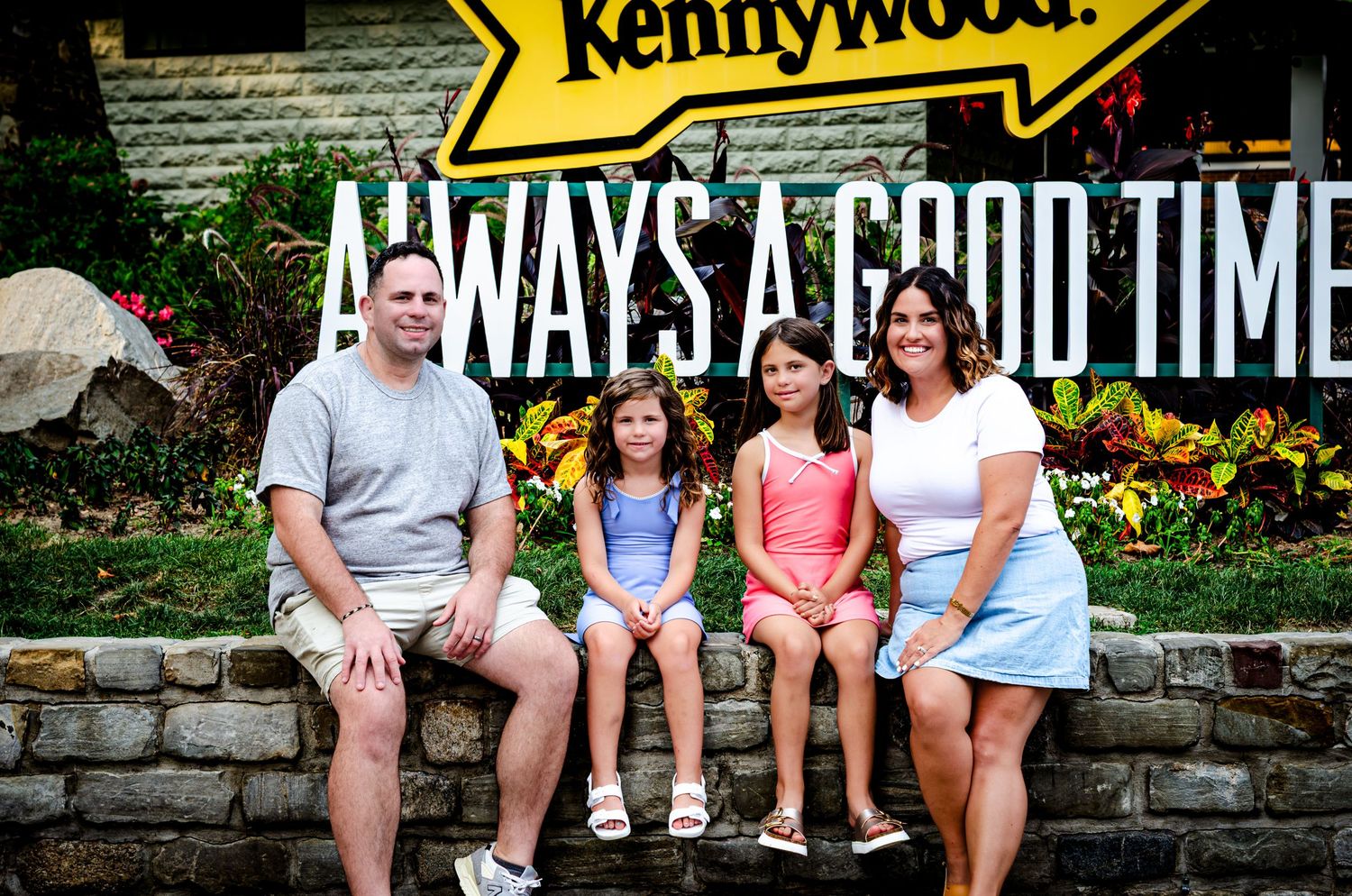 Family of four smiling in front of Kennywood amusement park Always a Good Time sign on a sunny day.
