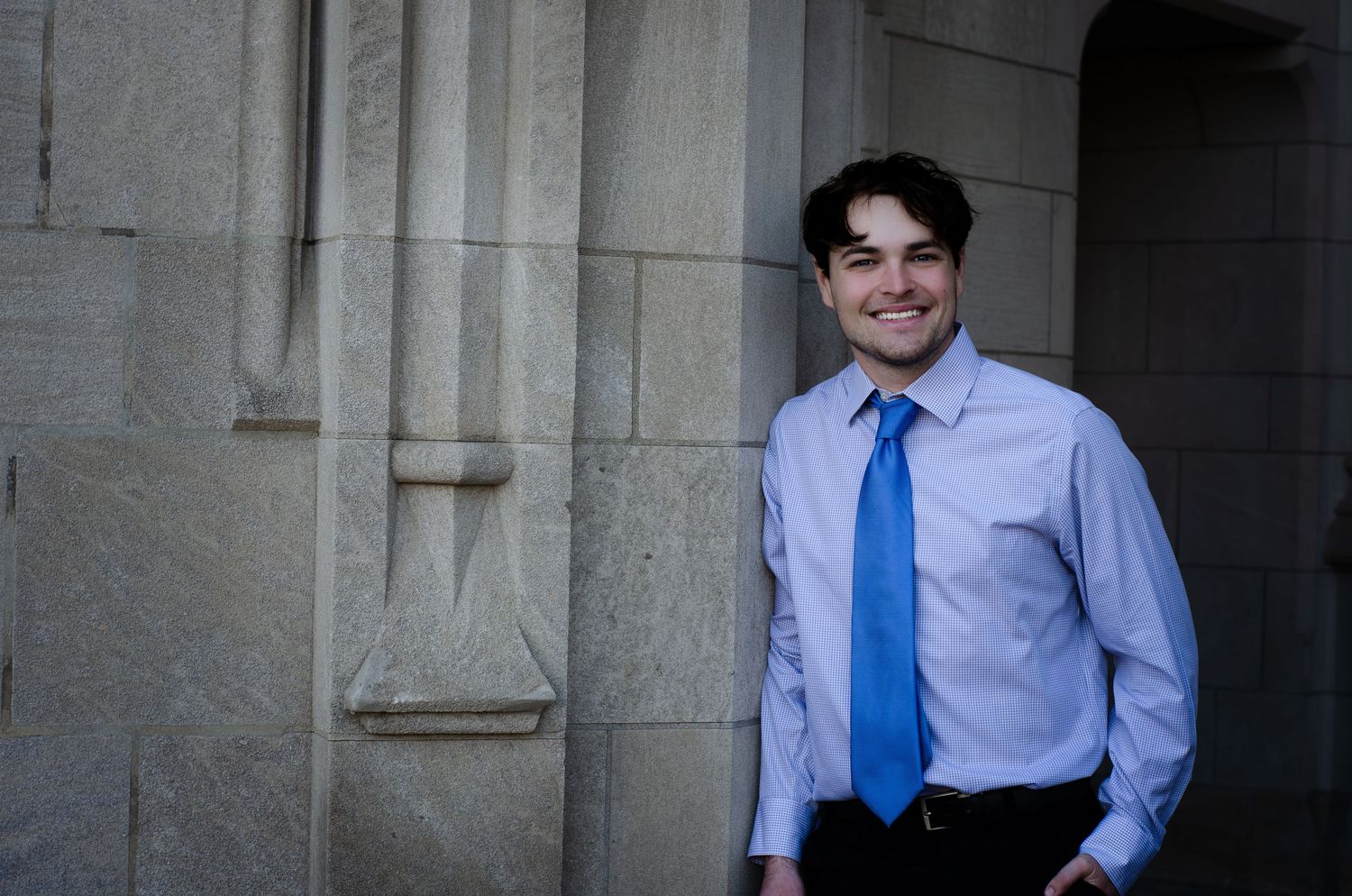 Smiling young man in blue striped shirt and tie leaning against stone building column.