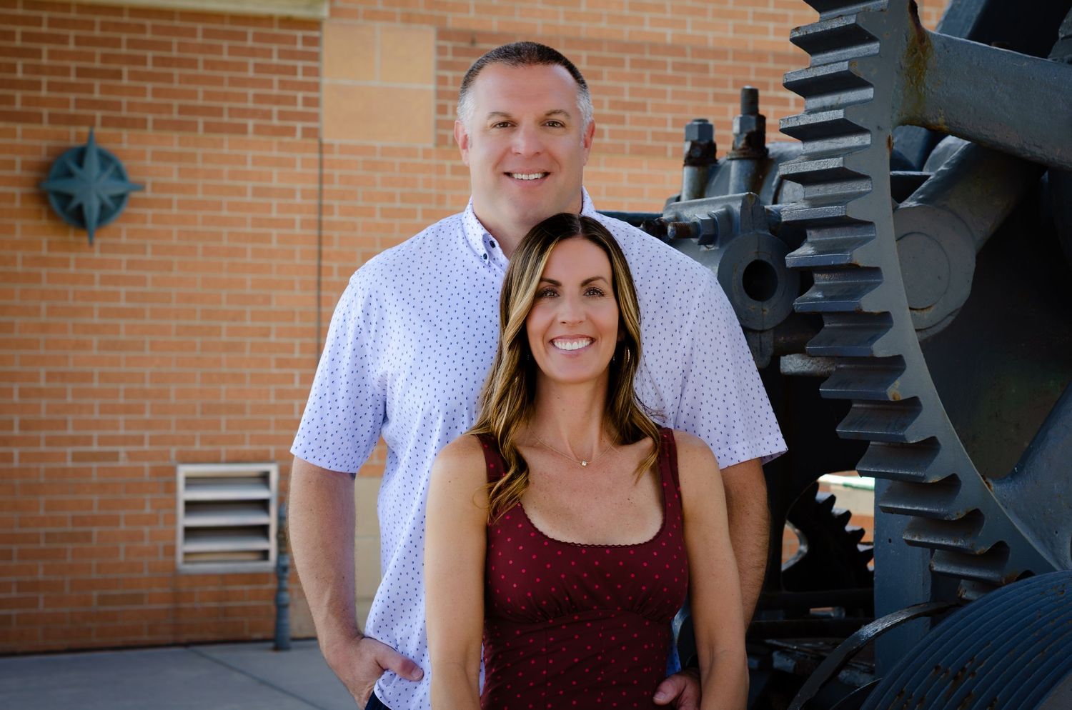 A smiling couple poses outdoors near large industrial gears and a brick wall on a sunny day.