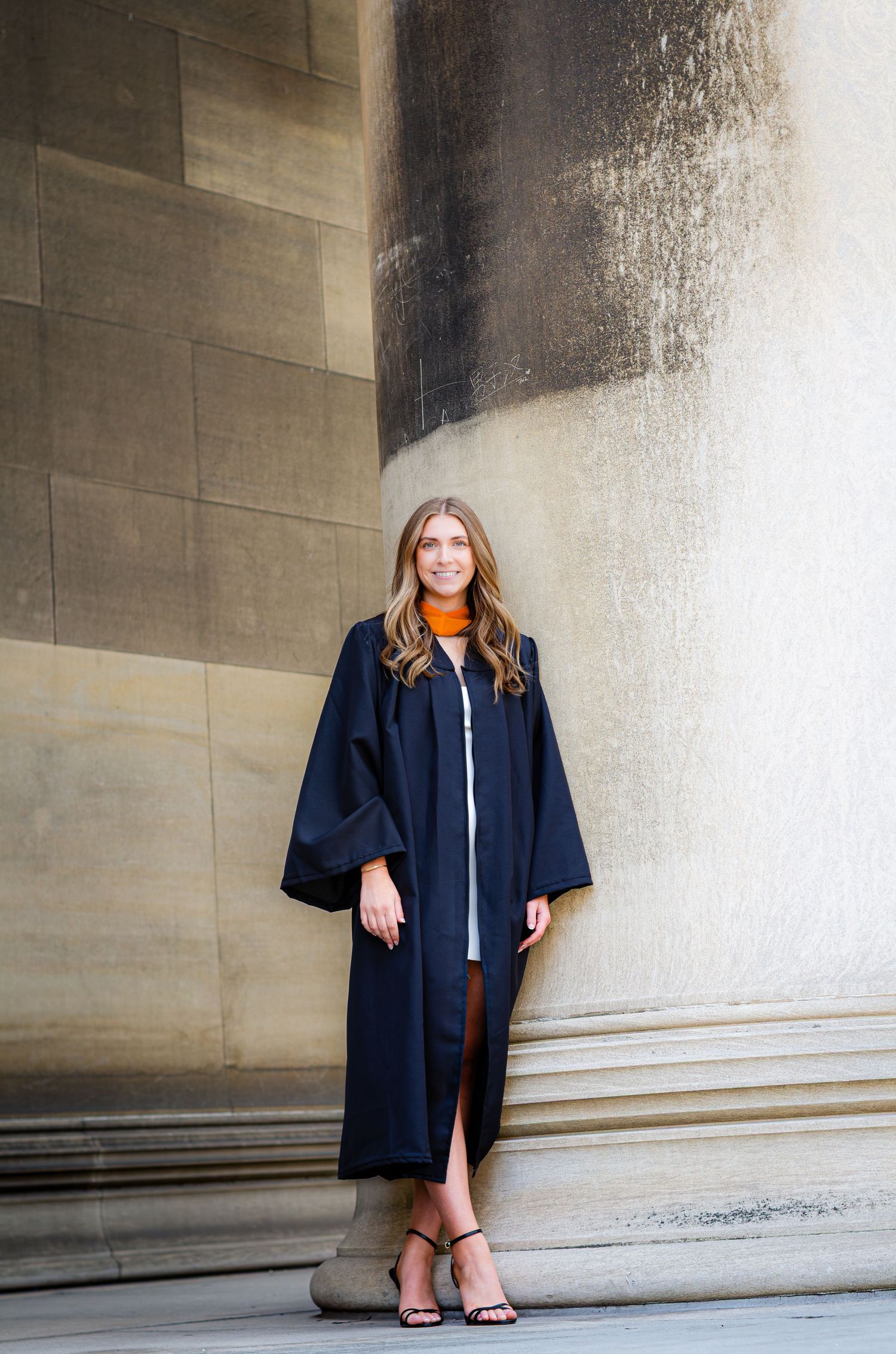 Graduate in navy academic gown with orange collar posing against large stone column.