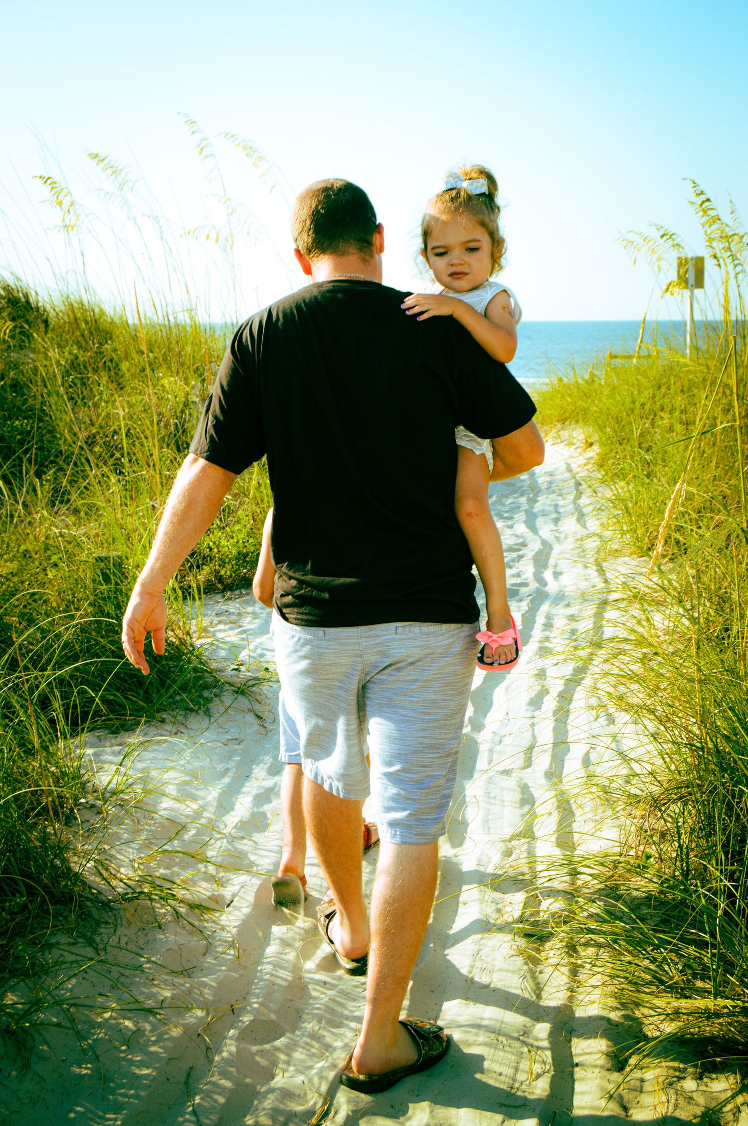 People walking down a sandy beach path through sea oats toward the ocean on a sunny day.