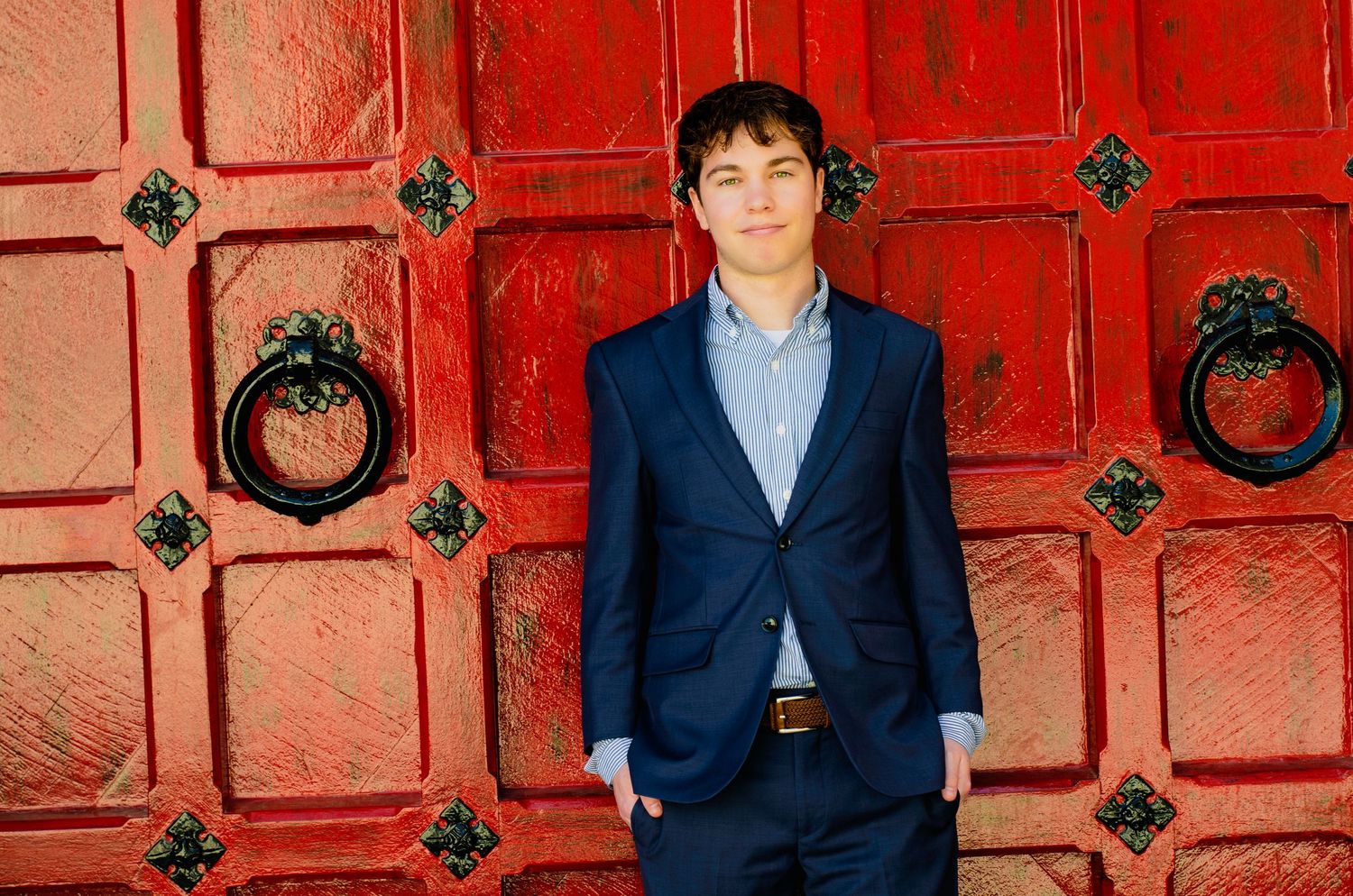 Young man in navy blue suit standing before ornate red double doors.