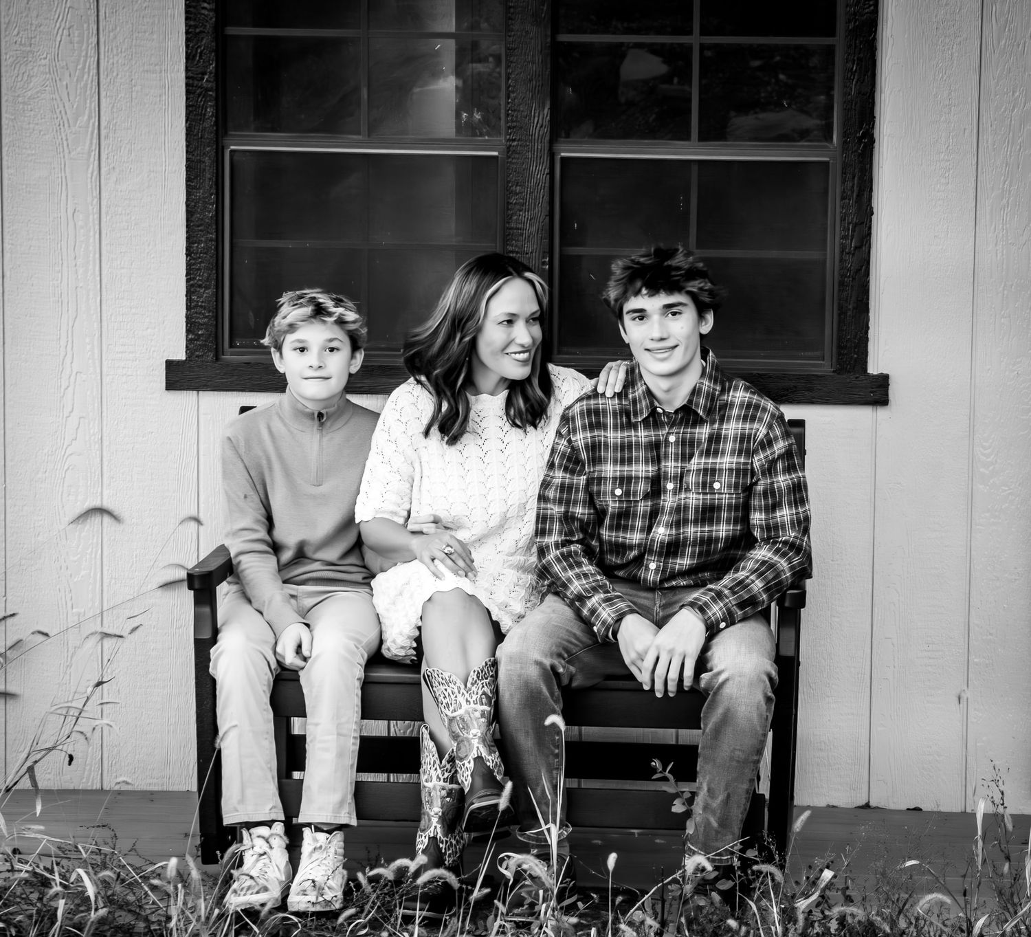 A black and white panoramic photo shows four people sitting casually on a porch ledge against a wooden wall.