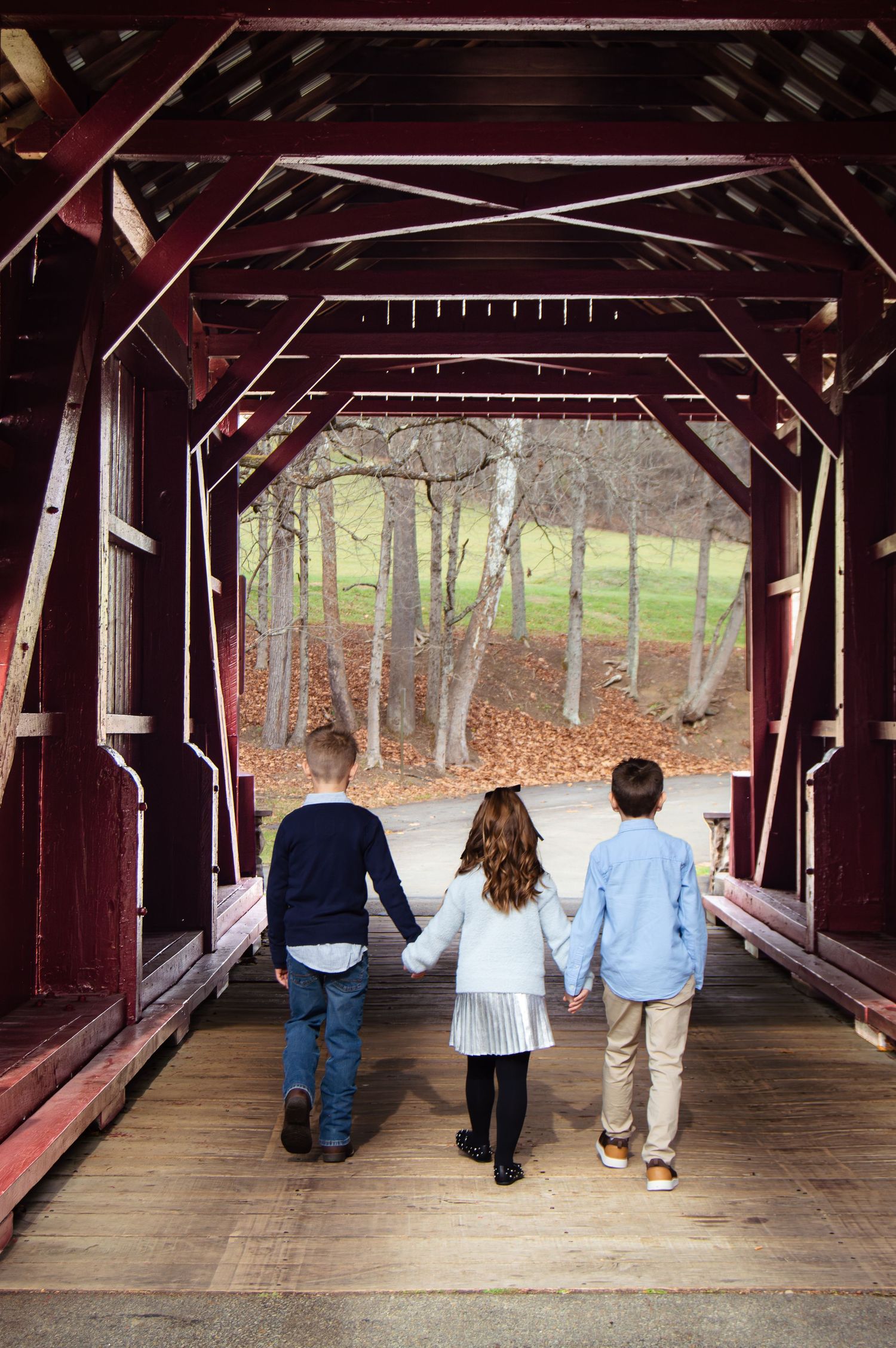 Three people walk together through a rustic covered wooden bridge looking out at autumn trees in the distance.
