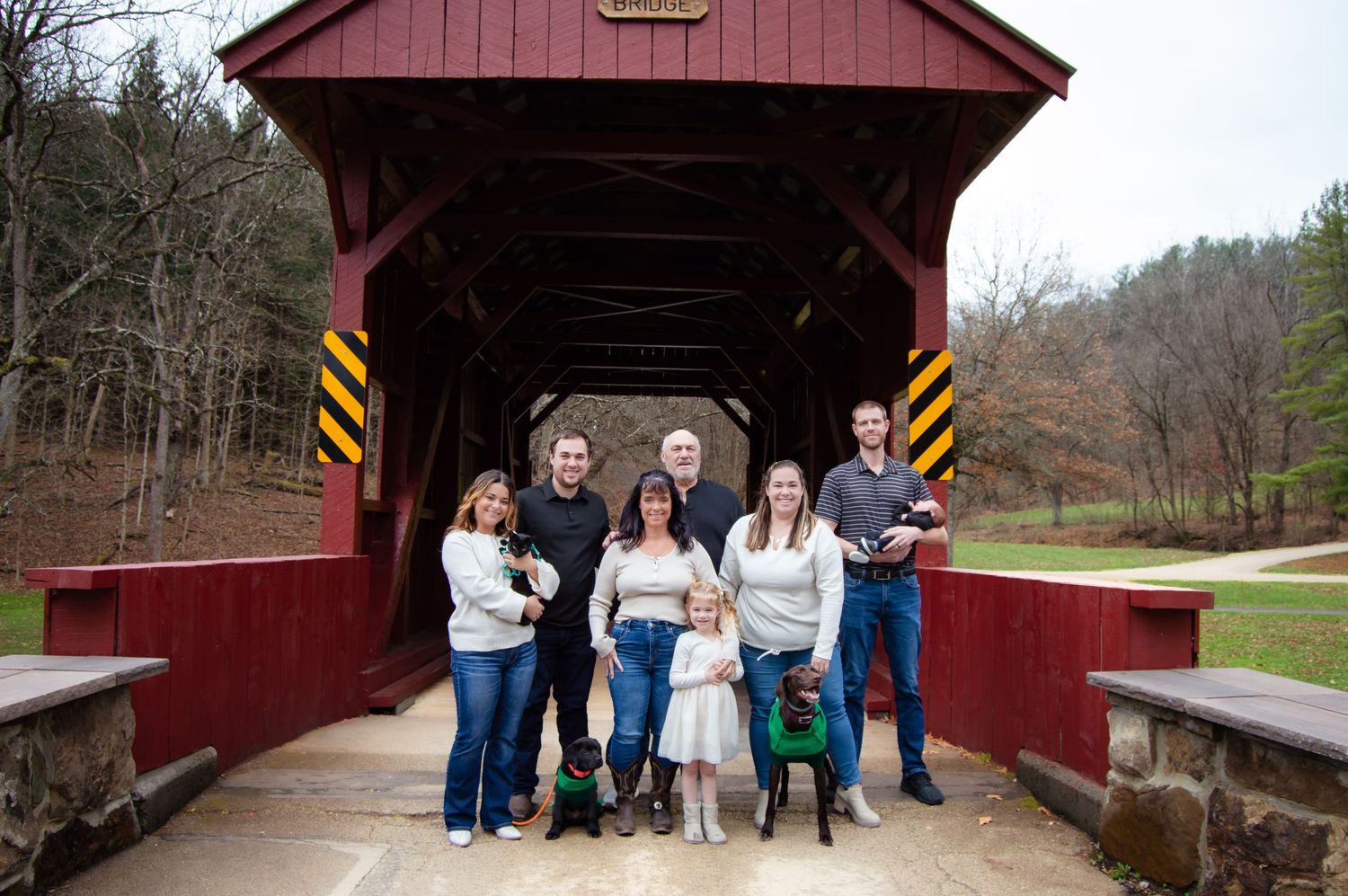 People gather for photos at a rustic red covered bridge on a cloudy autumn day.