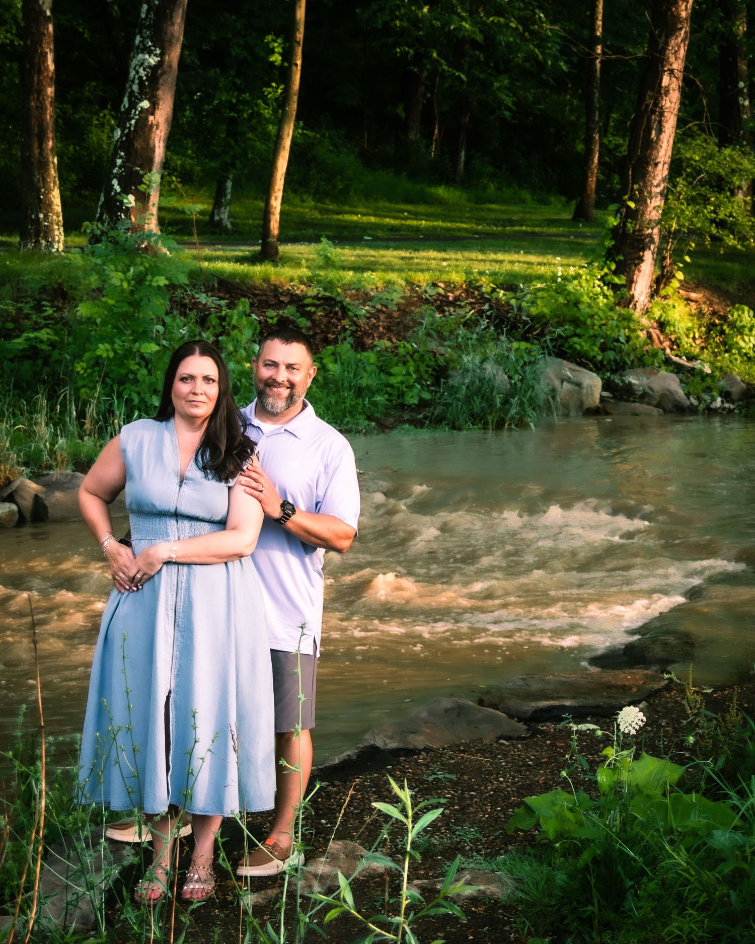 A couple in light blue and white outfits poses together by a wooded creek bank in summer.