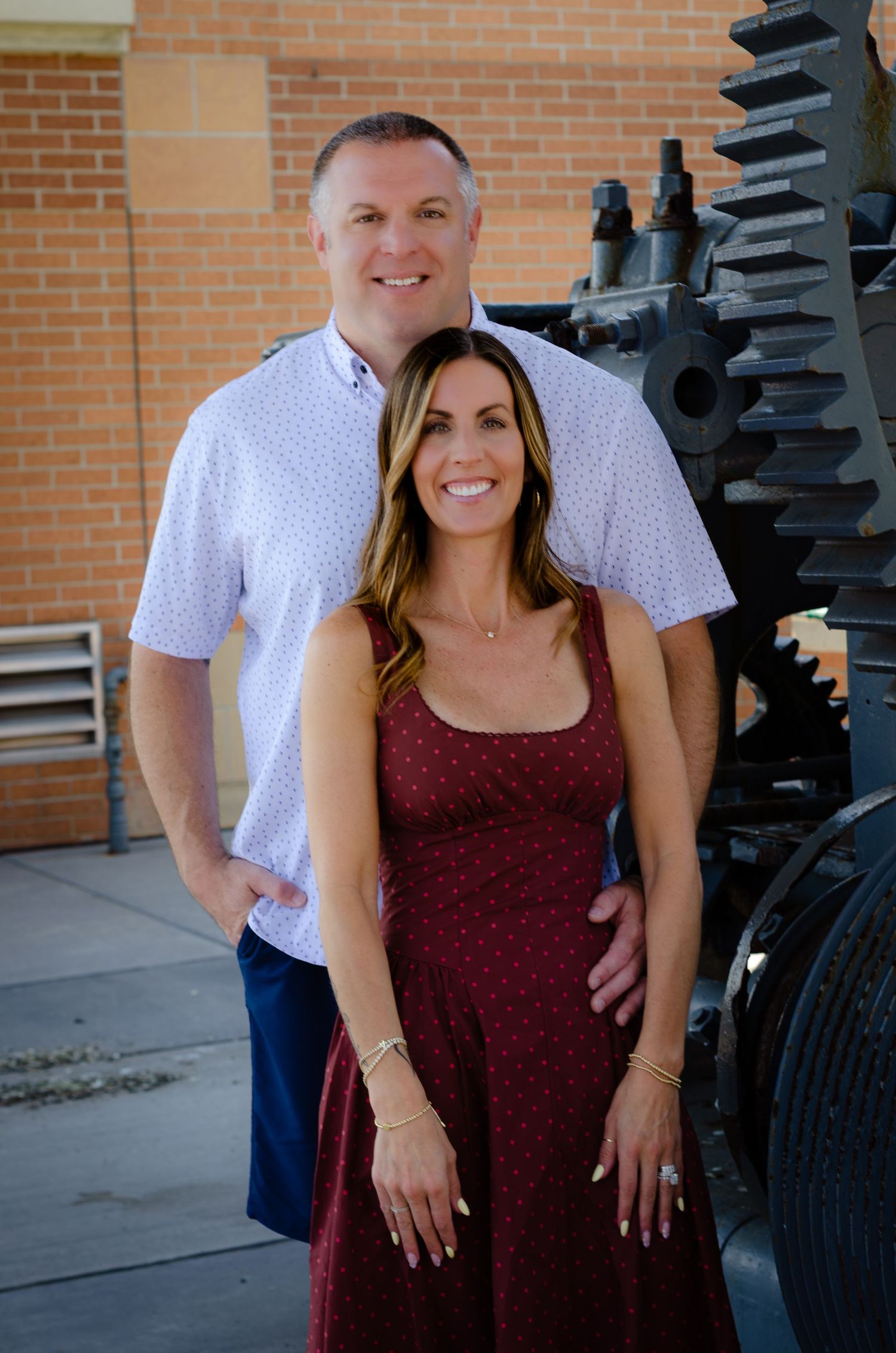 Smiling couple posing outdoors near vintage industrial gears beside a brick building.