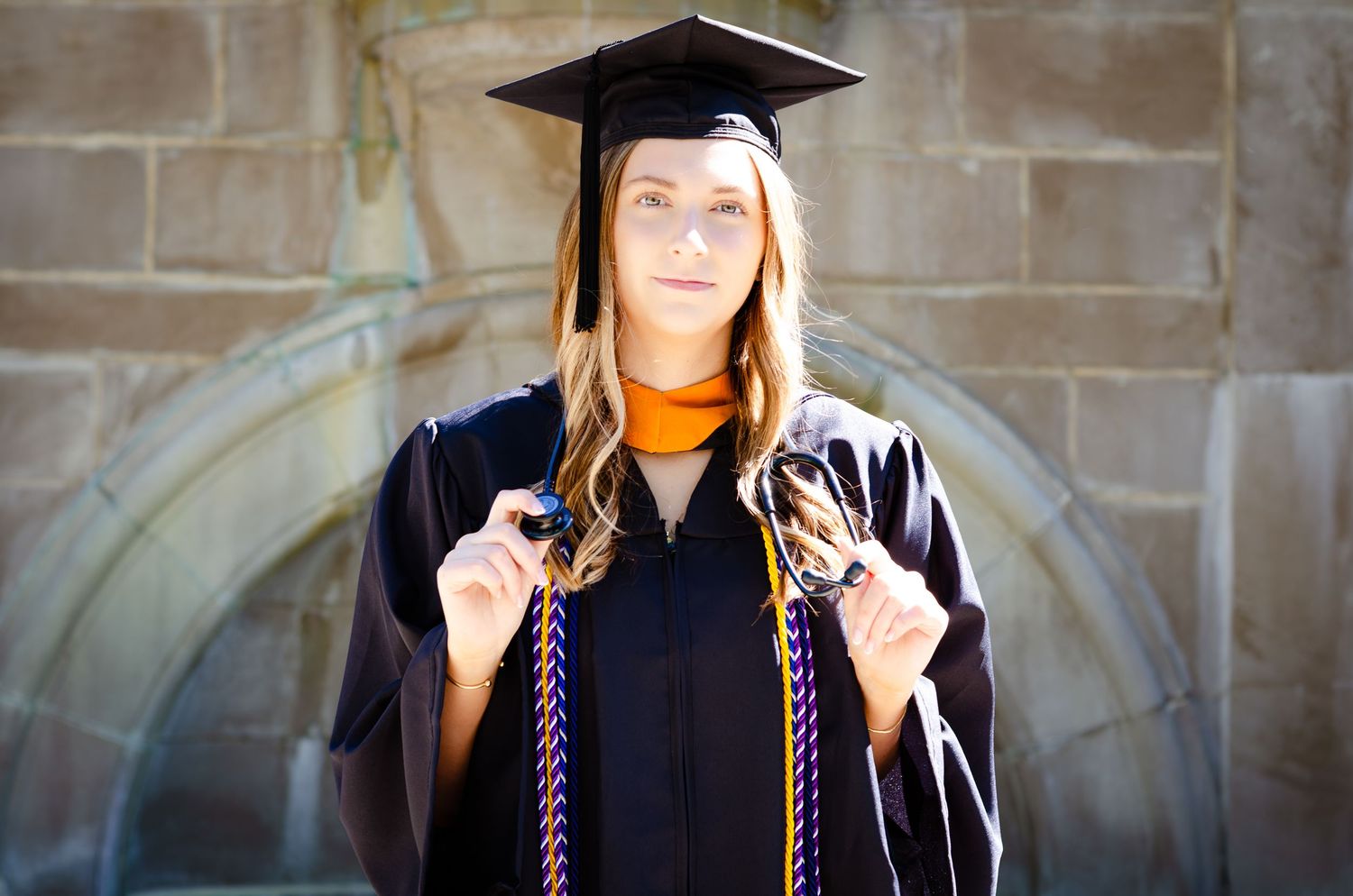 Young woman in black graduation cap and gown with colorful honor cords posing in front of stone building.