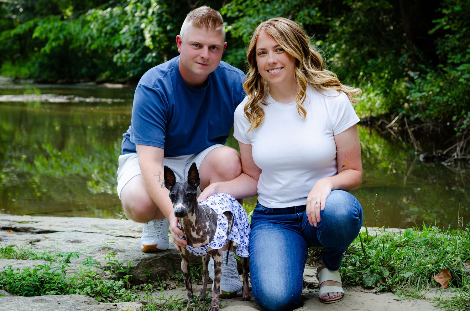 Couple crouching with their dog in a spotted outfit near a creek surrounded by lush greenery.