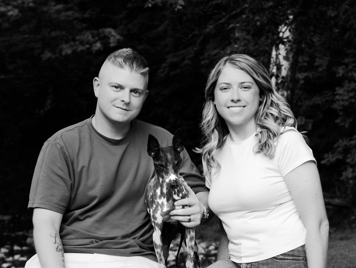 Black and white photo of a smiling couple posing outdoors with their spotted dog among trees.