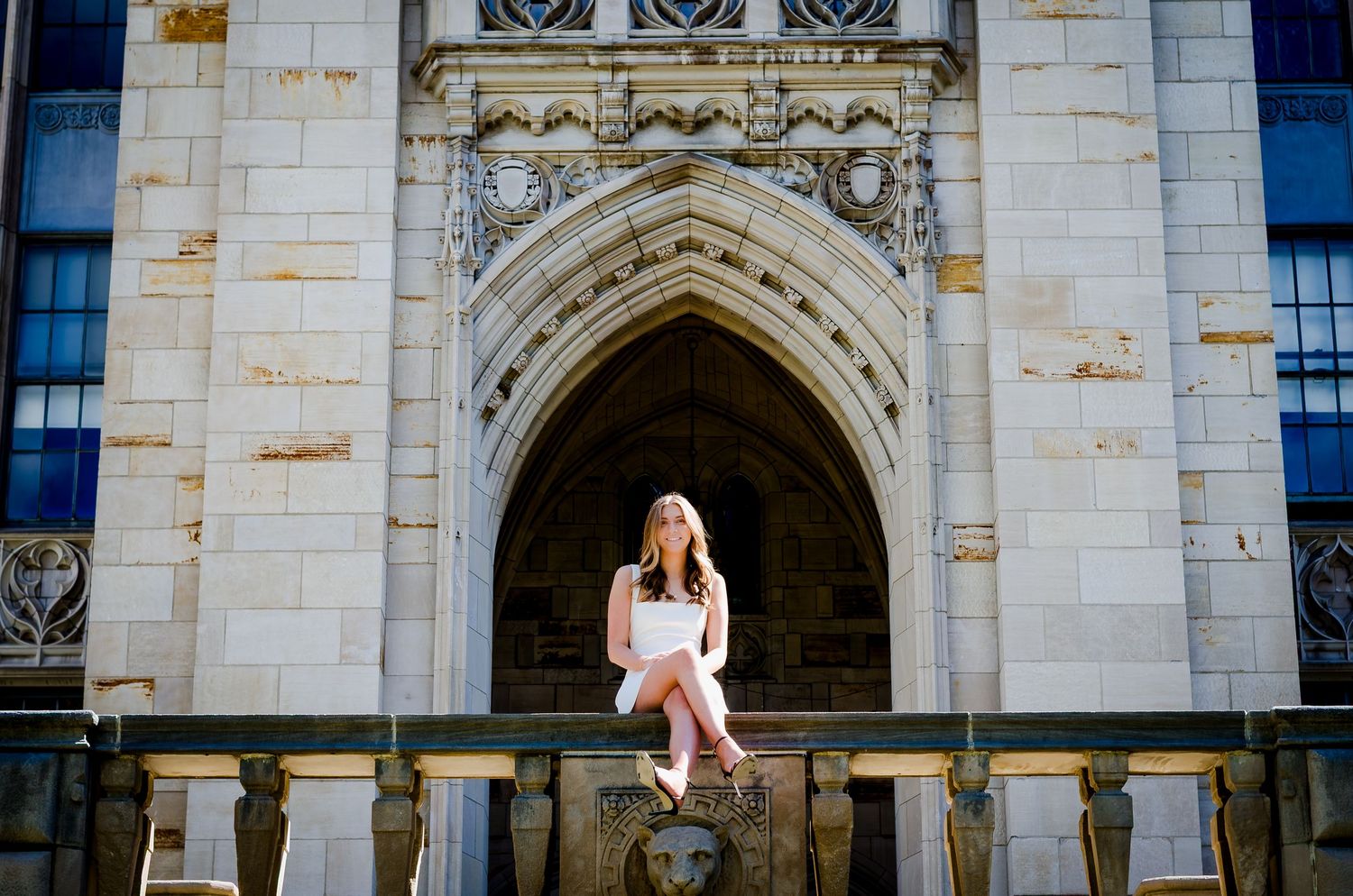 Young woman in white dress sitting on stone ledge beneath ornate Gothic arch of university building.