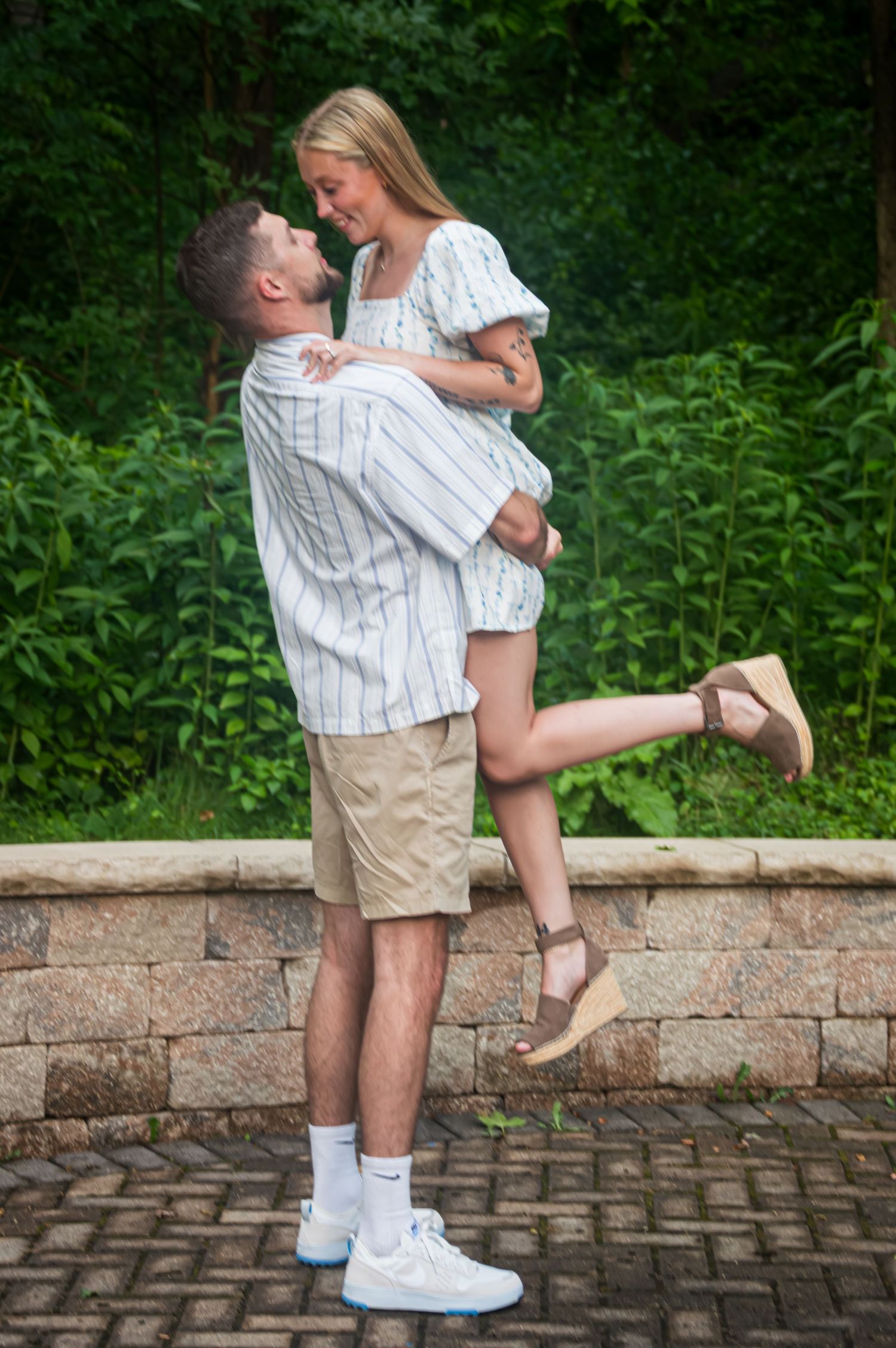 Romantic couple in white and khaki embracing by a stone wall in a garden setting.