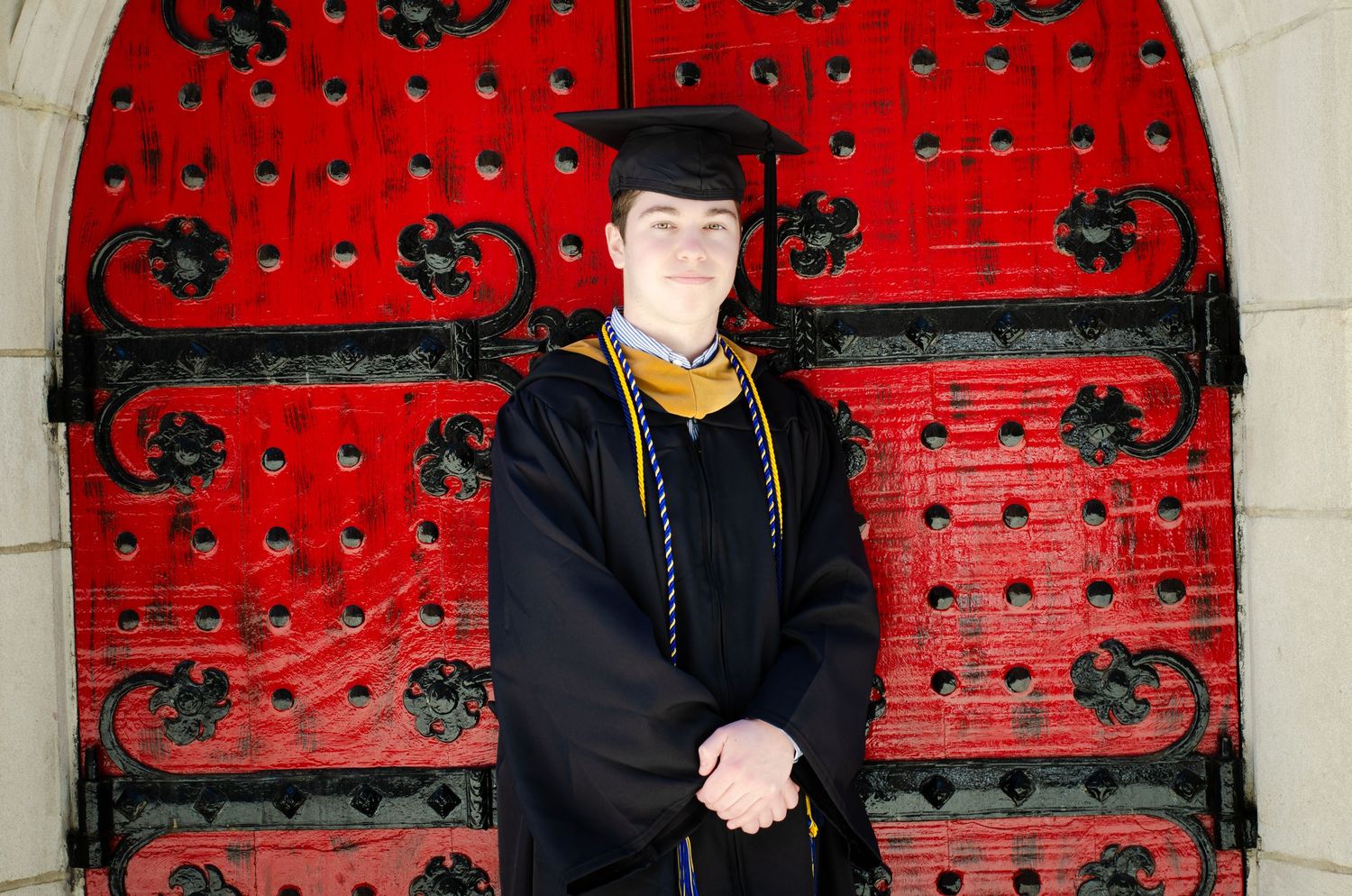 Graduate in black cap and gown with gold stole posing before red ornate doors.