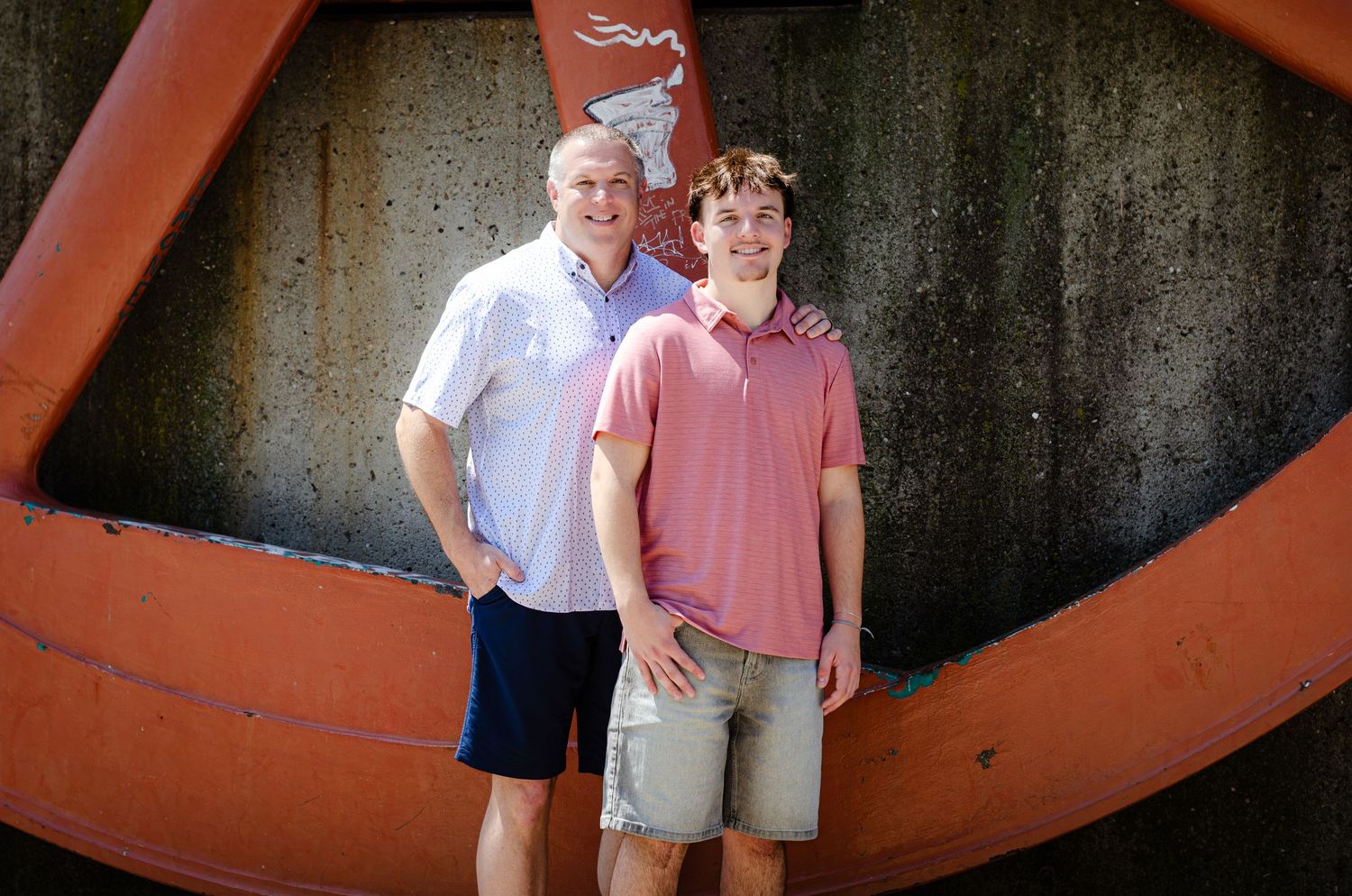 Man and teenage boy smiling together outdoors in front of large red decorative wheel structure.