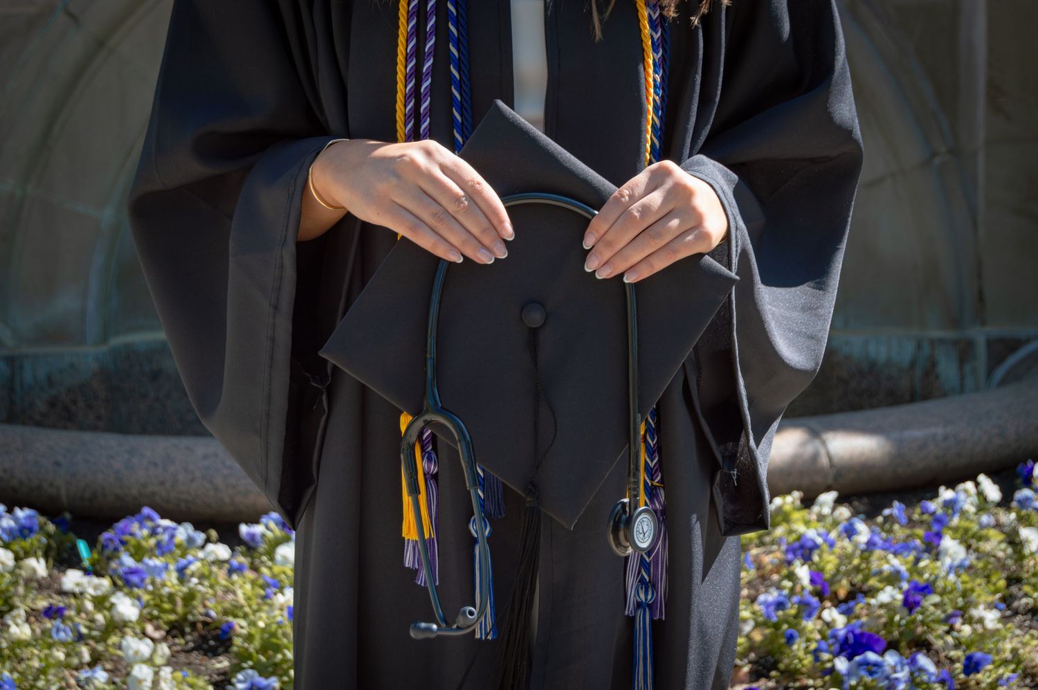 Graduate in black cap and gown holding mortarboard at outdoor ceremony with blue flowers in background.