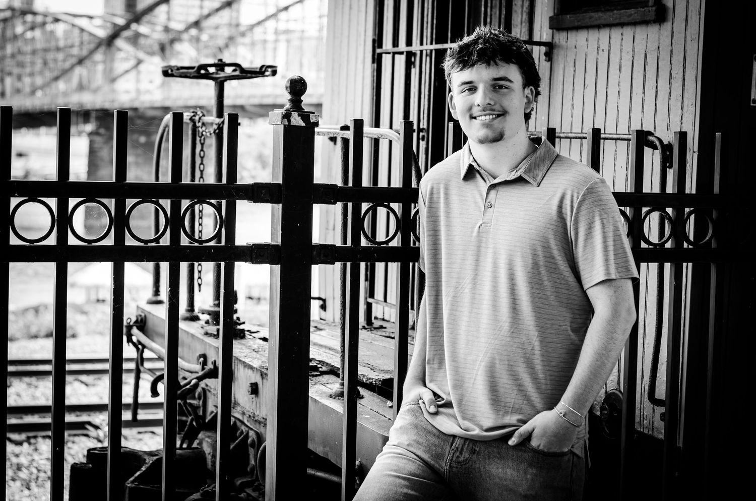 Young man smiling in black and white portrait, leaning against ornate iron fence in urban setting.