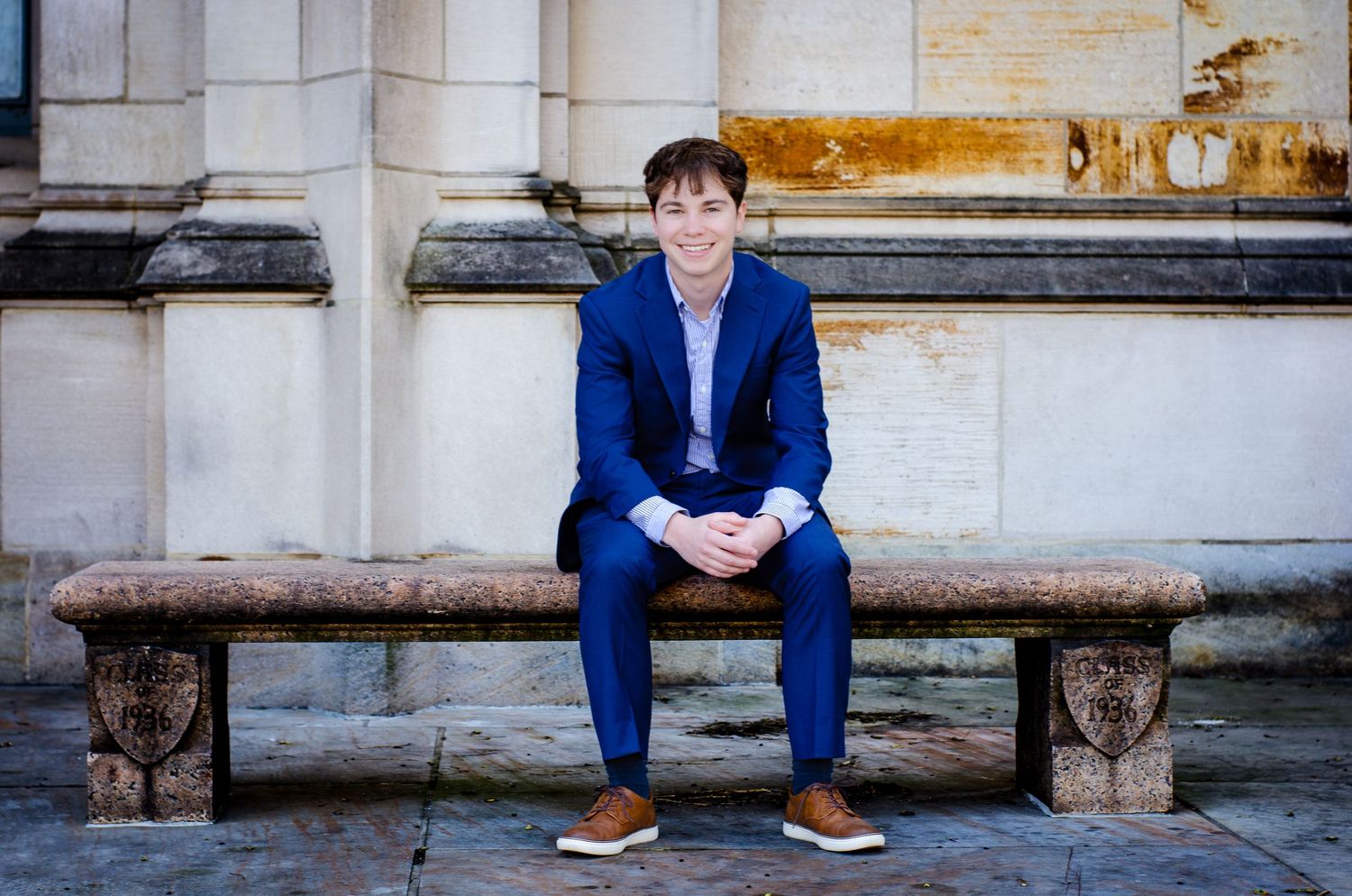 Young man in blue suit sitting on stone bench outdoors, smiling confidently in front of a classic building.