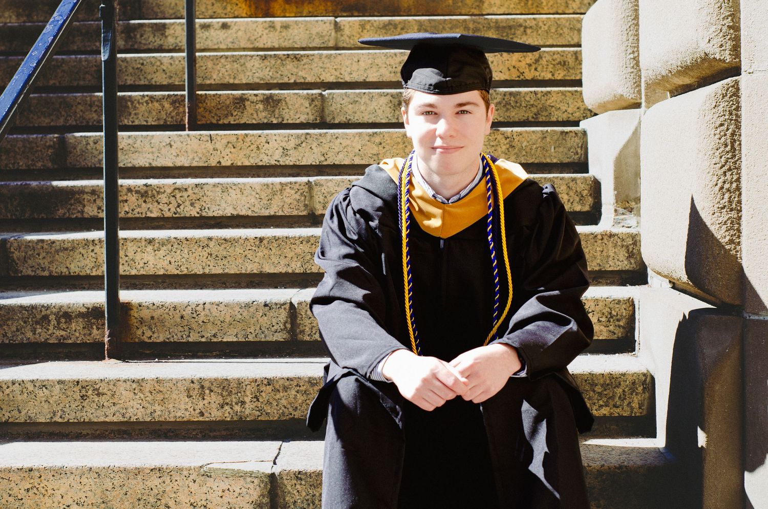 Graduate in black cap and gown with gold honors cords sits on stone steps, smiling for graduation photo.